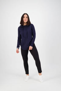 A woman with shoulder-length dark hair stands against a white background, wearing Vassalli's Fortune Pant in black, a navy hoodie, and white sneakers. She smiles slightly with a relaxed posture.