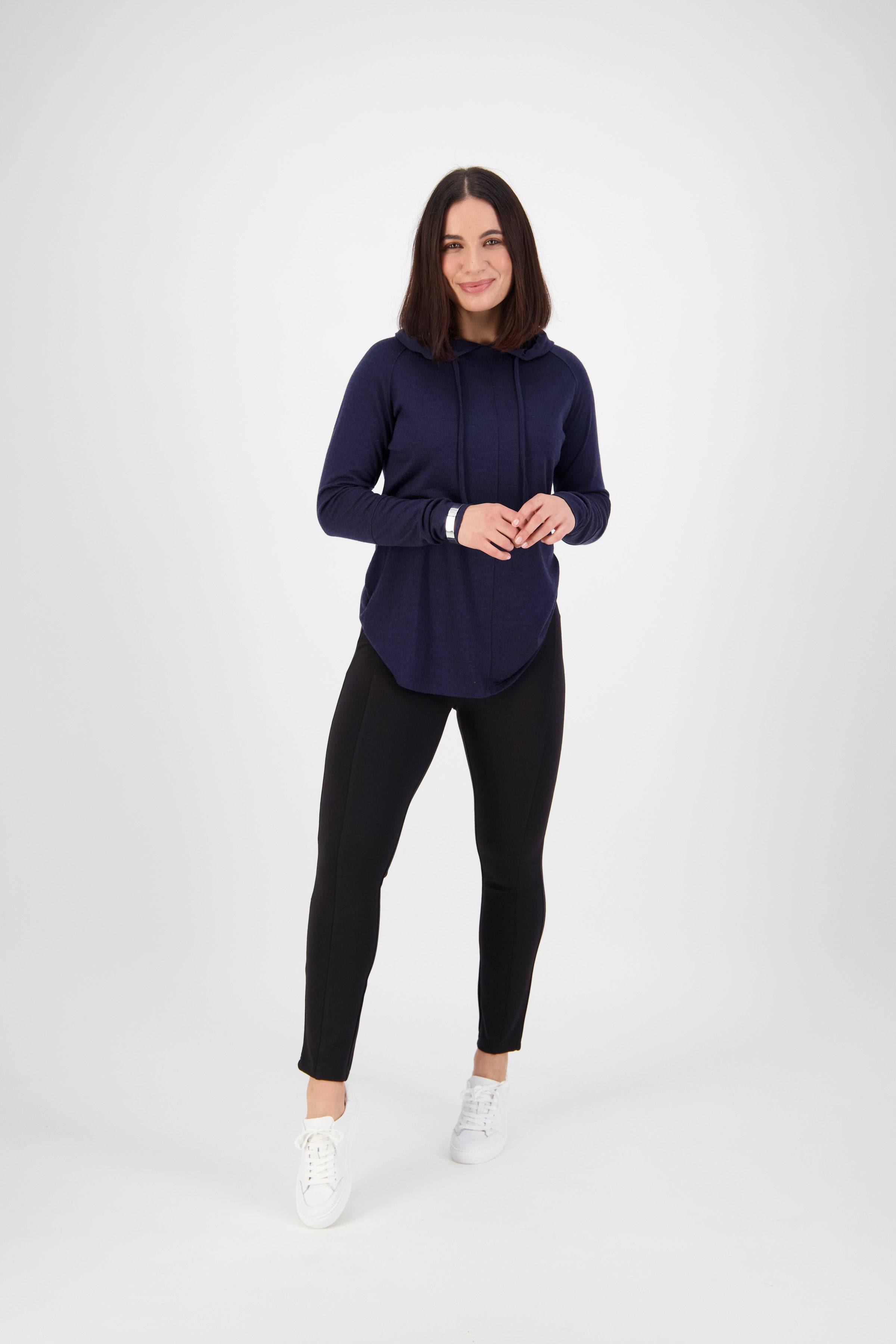 A woman with shoulder-length dark hair smiles, wearing a navy hoodie, Vassalli Fortune Pant - Black, and white sneakers against a plain white background.