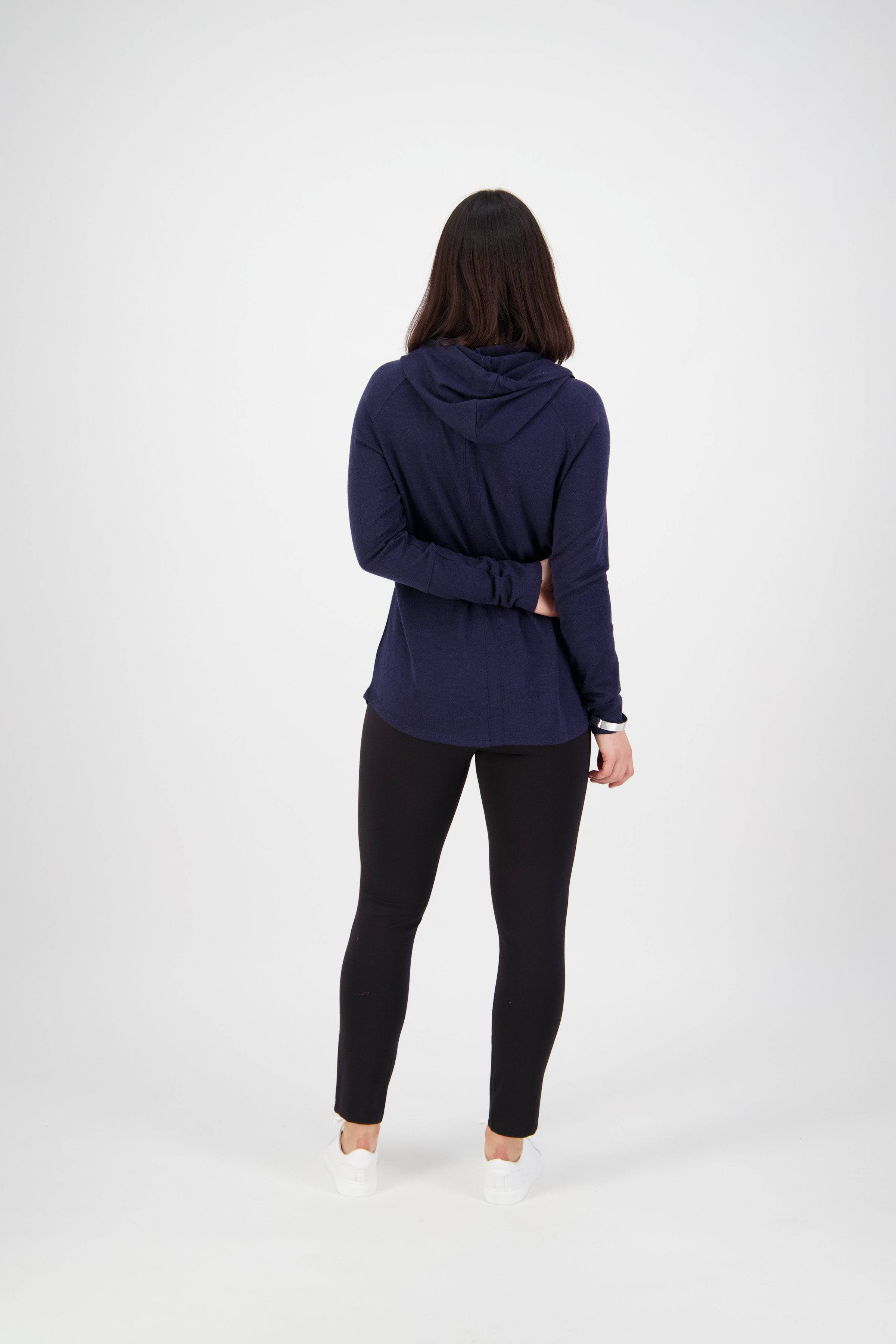 A person with shoulder-length dark hair faces away, wearing a navy hooded sweatshirt and Vassalli Fortune Pant - Black. Their right arm is bent behind their back, holding their left elbow. The background is plain white.