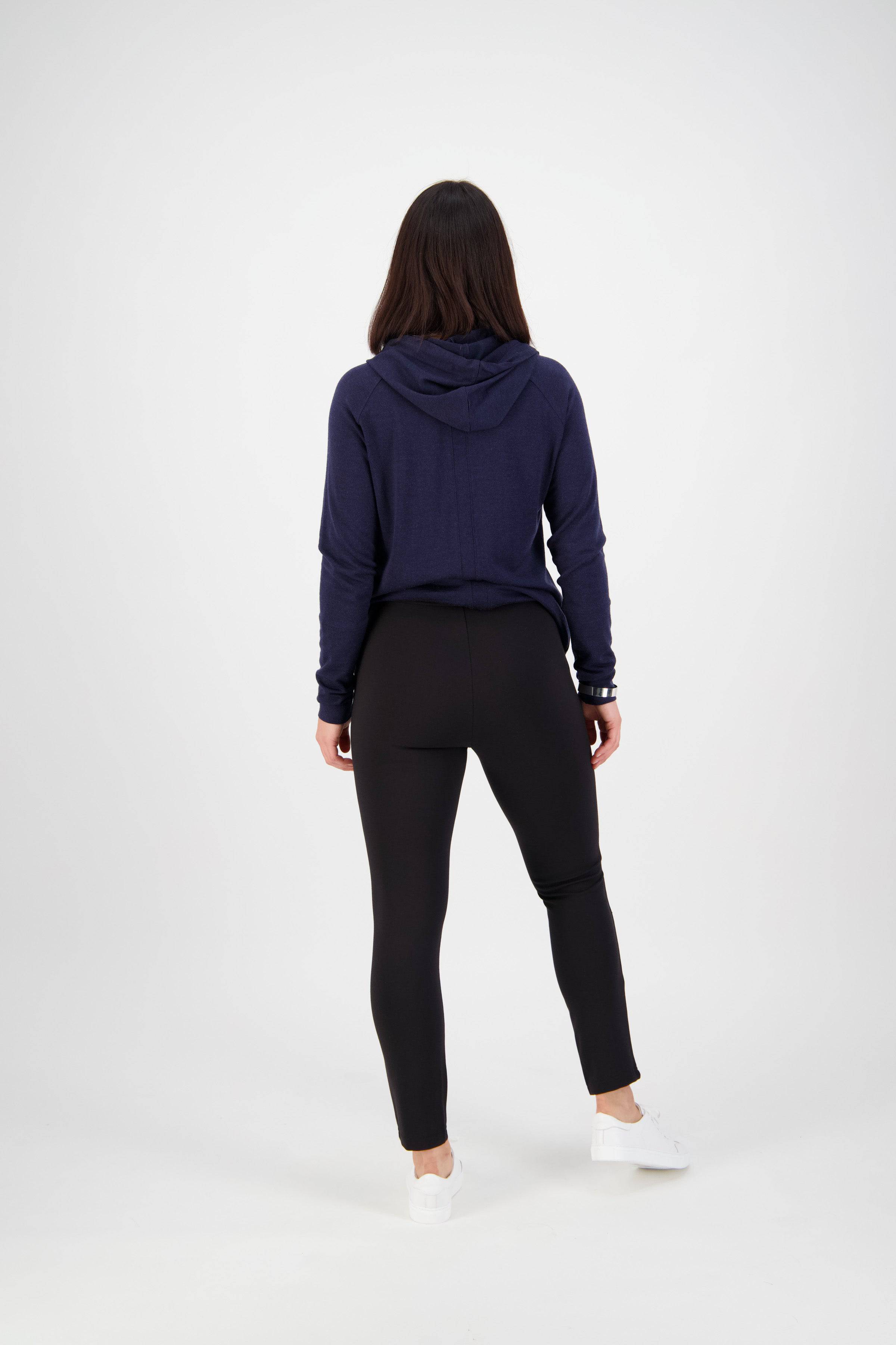 A person with long dark hair faces away, wearing a navy blue hoodie, Vassalli Fortune Pant in black, and white sneakers against a plain white background.