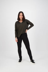 A woman with shoulder-length dark hair stands confidently against a plain white background, wearing a dark green long-sleeve top, Vassalli Toll Pant in Black, and black sneakers, one hand on her hip and a slight smile.