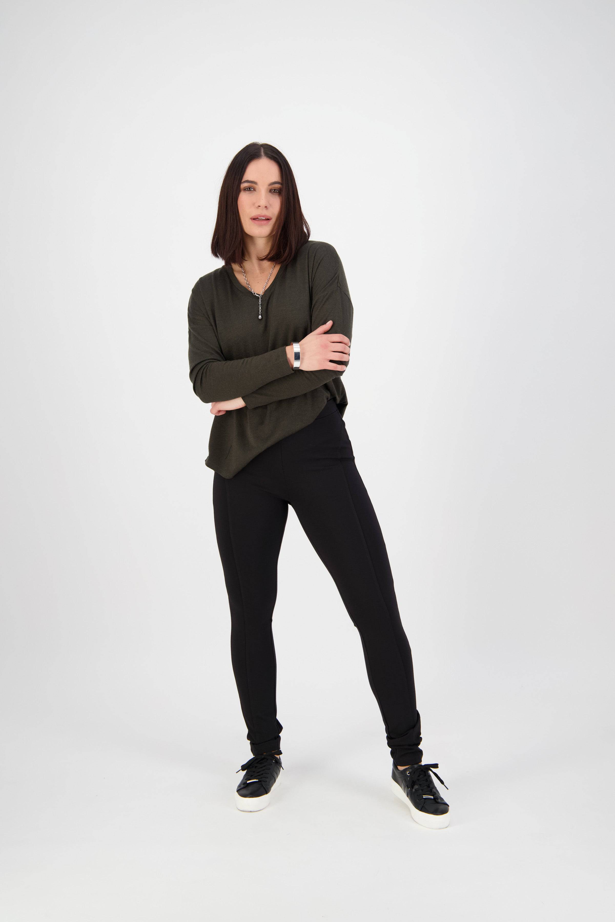A woman with straight dark hair stands confidently with arms crossed, wearing a green long-sleeve top, Vassalli Toll Pant - Black, black sneakers, and a smart watch against a plain white background.