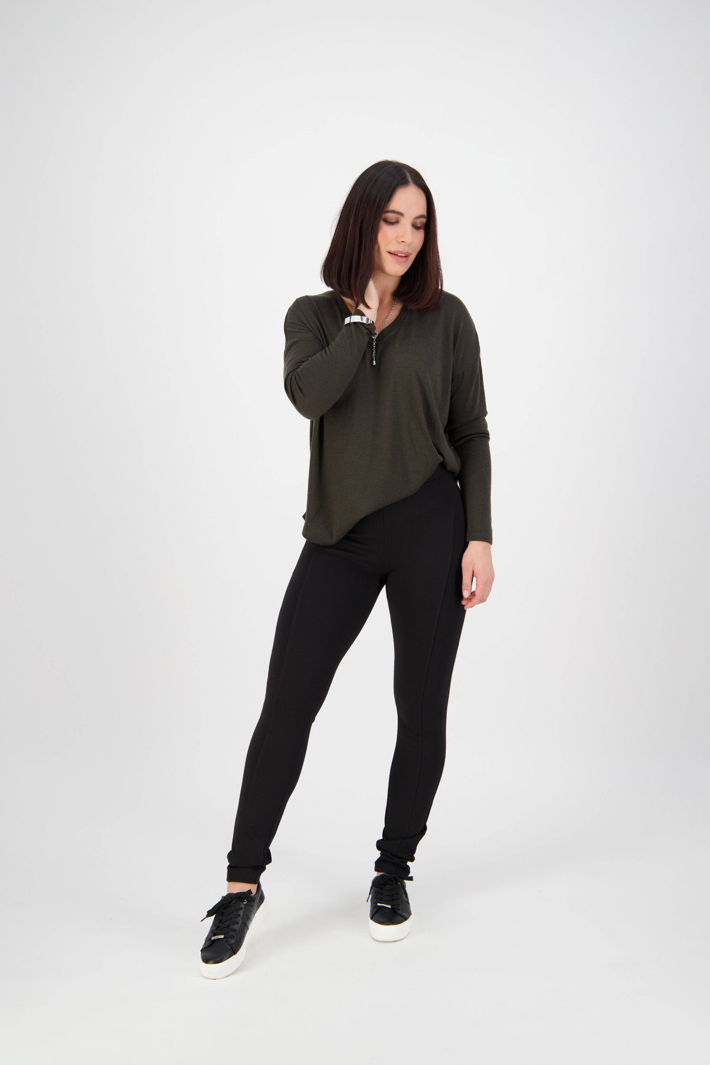 A woman with straight, shoulder-length dark hair stands against a plain white background. She wears a loose-fitting dark green top and Vassalli Toll Pant in black, a pull-on skinny leg style made from stretch fabric, with one hand touching her neck.