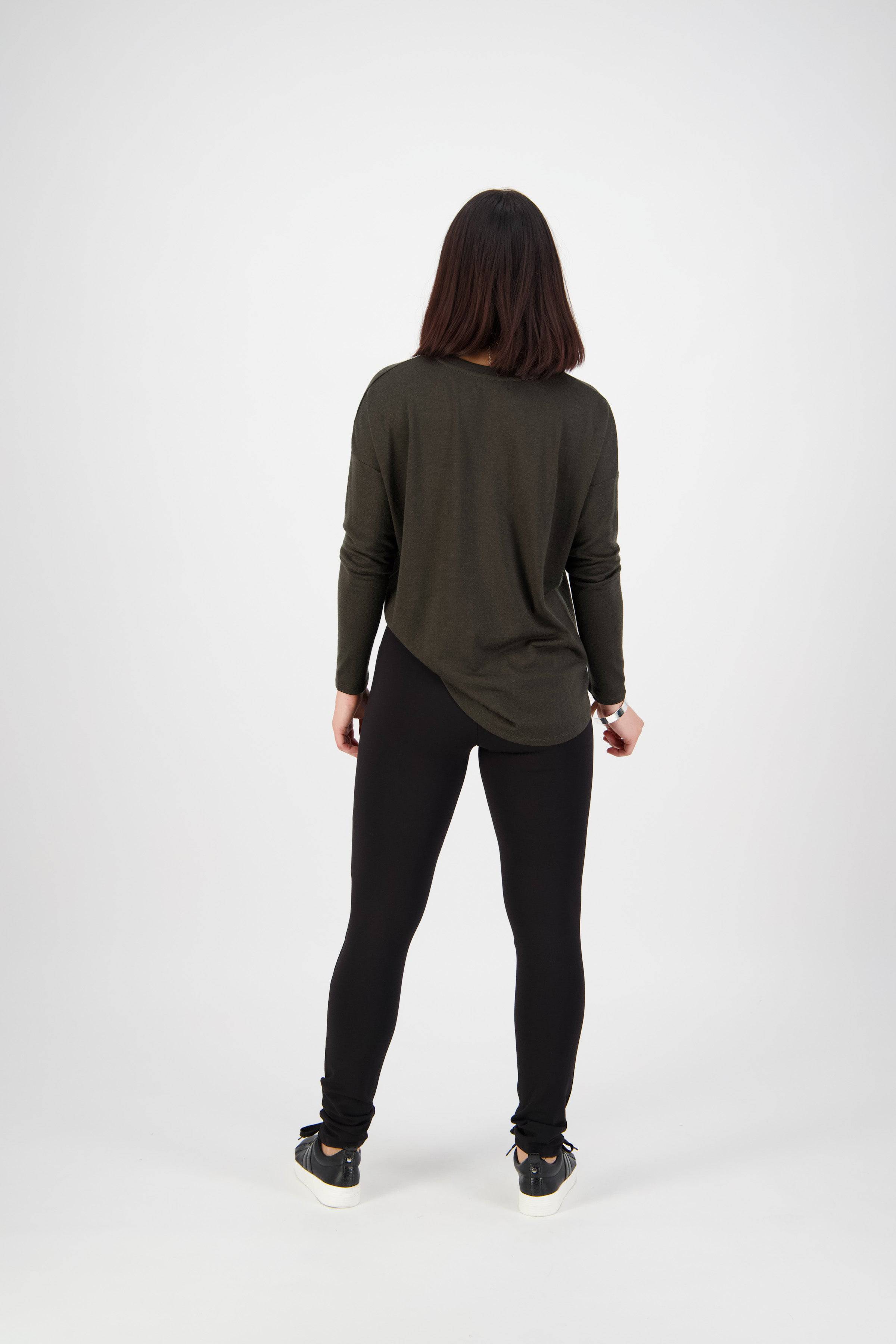 A woman faces away in a studio, wearing the Vassalli Toll Pant - Black, styled with a long-sleeve dark green top and black sneakers with white soles. The backdrop is plain and white.