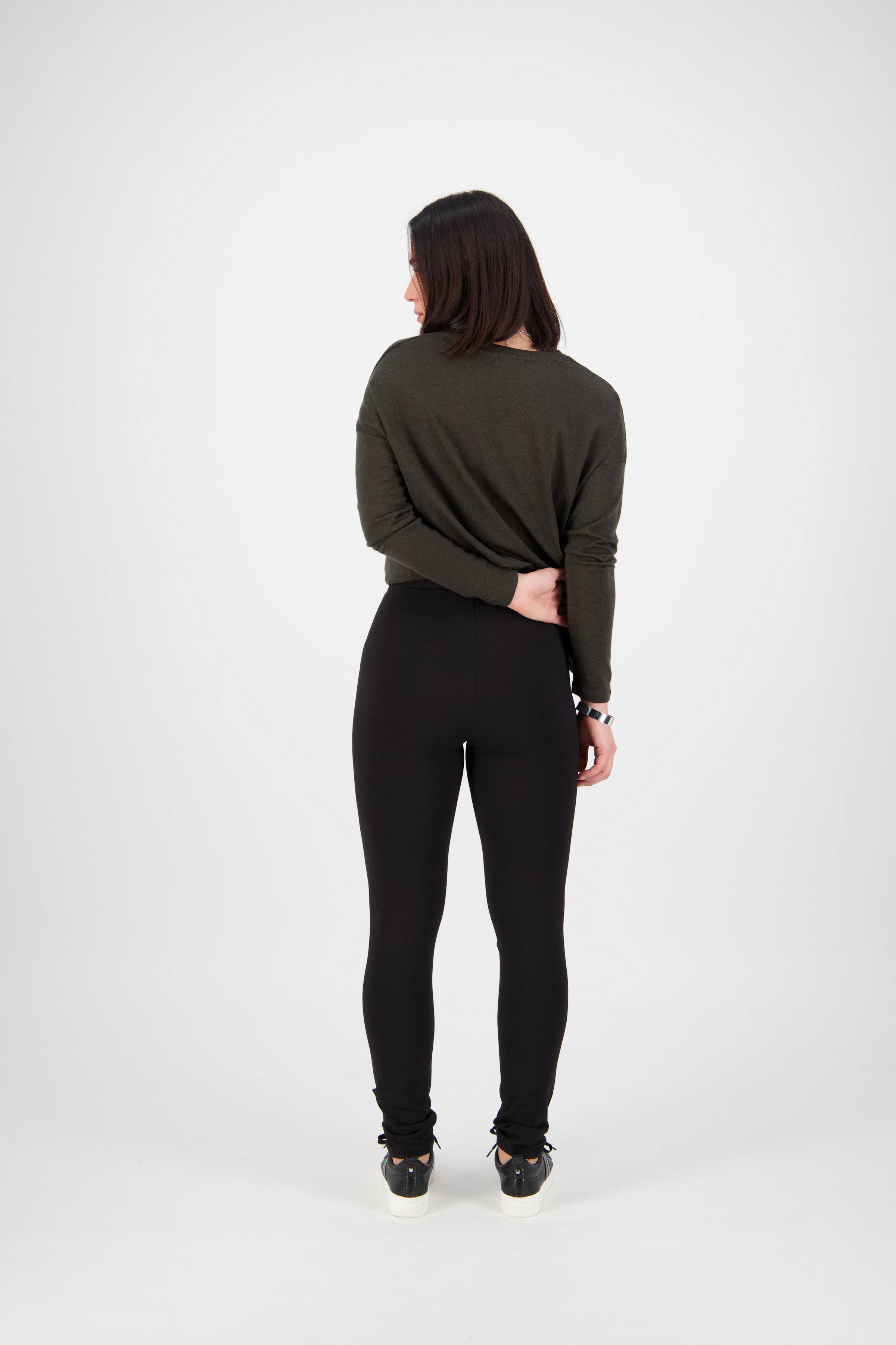 A woman with straight dark hair faces away, wearing a dark green long-sleeve top and Vassalli’s Toll Pant - Black skinny pull-on pants, made from stretch fabric, with one wrist held behind her back against a white background.