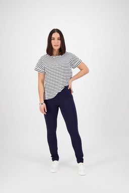 A woman with shoulder-length dark hair poses against a white background wearing the Vassalli Toll Pant in navy, a black and white striped short-sleeve top, and white sneakers, with one hand confidently on her hip.