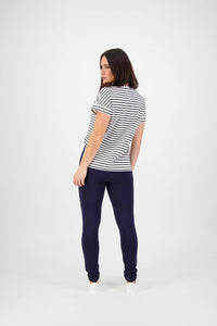 A woman with shoulder-length dark hair stands slightly turned, wearing a short-sleeved white shirt with black stripes and Vassalli Toll Pant - Navy, made from stretch fabric, against a plain white background.