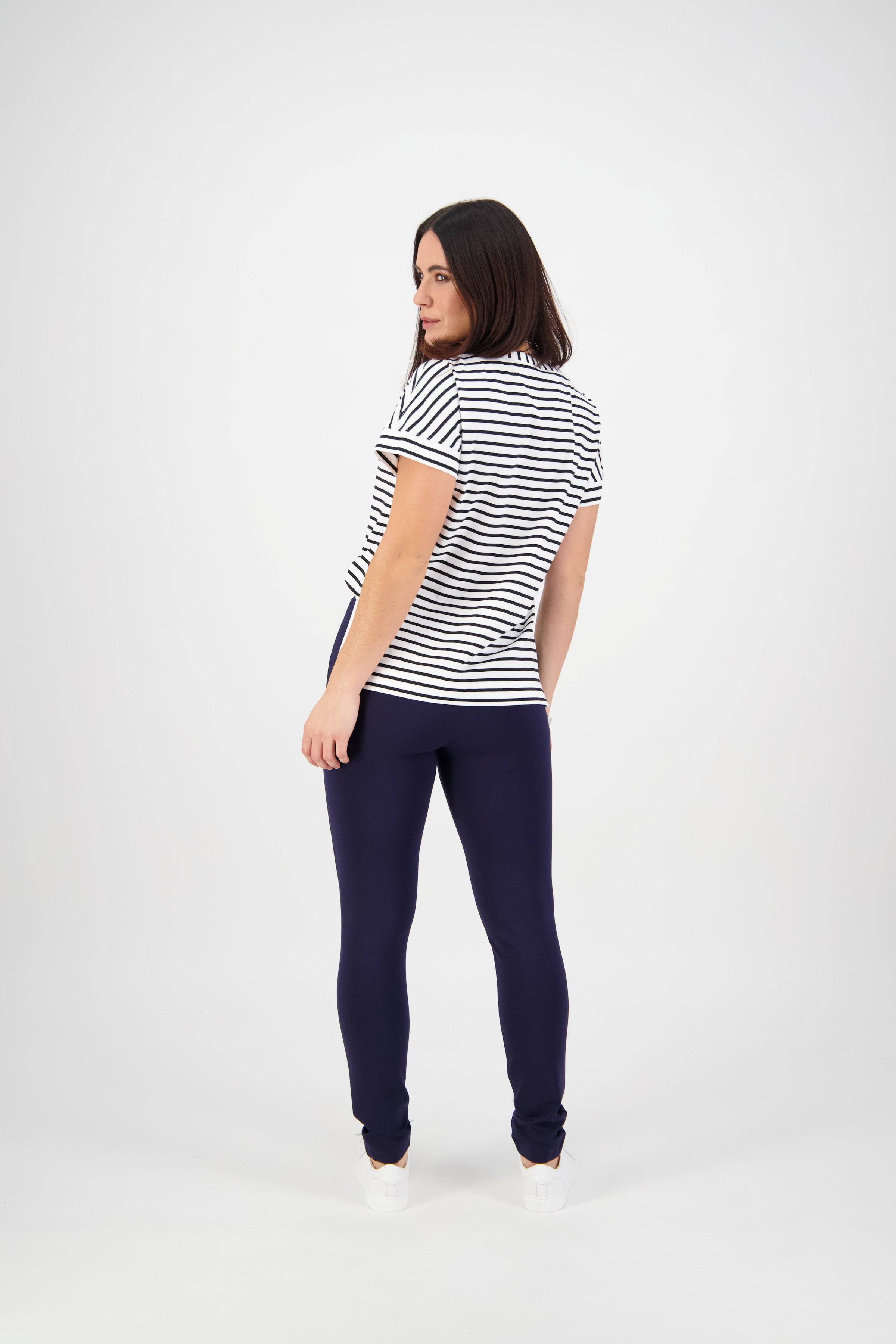 A woman with shoulder-length dark hair stands slightly turned, wearing a short-sleeved white shirt with black stripes and Vassalli Toll Pant - Navy, made from stretch fabric, against a plain white background.