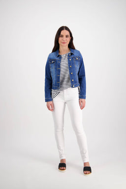 A woman with long dark hair and a relaxed expression wears the Vassalli Ratio Jacket in Blue Wash Denim over a black-and-white striped shirt, paired with white pants and black sandals, standing against a plain white background.