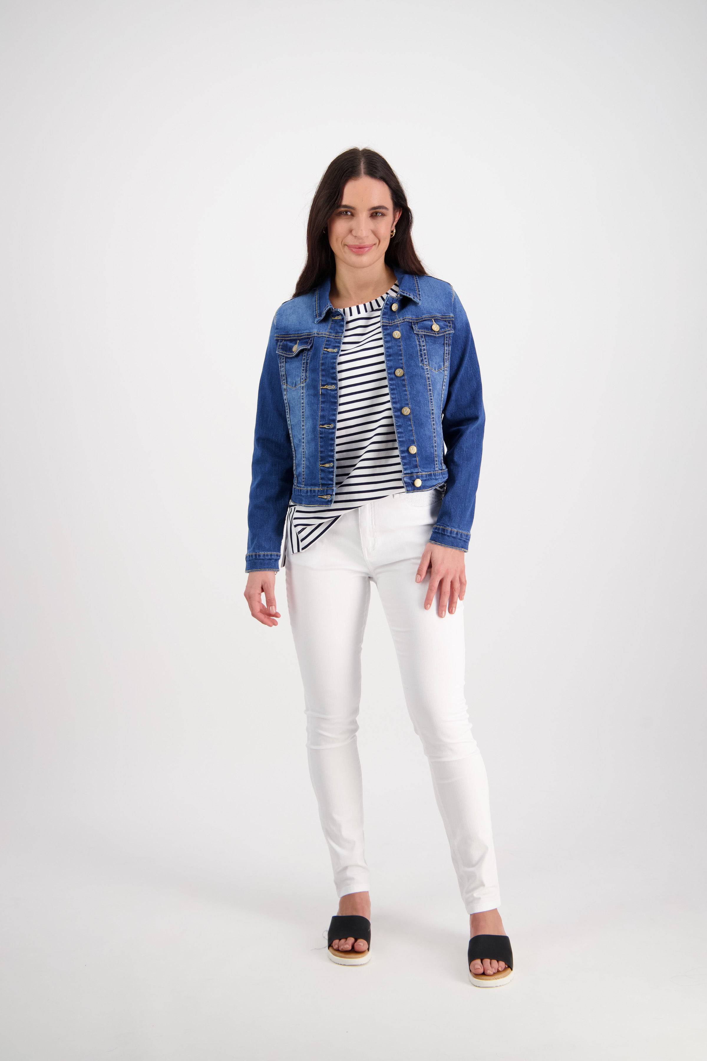 A woman with long dark hair smiles against a white background, wearing the Vassalli Ratio Jacket in Blue Wash Denim, paired with a black and white striped top, white pants, and black sandals.