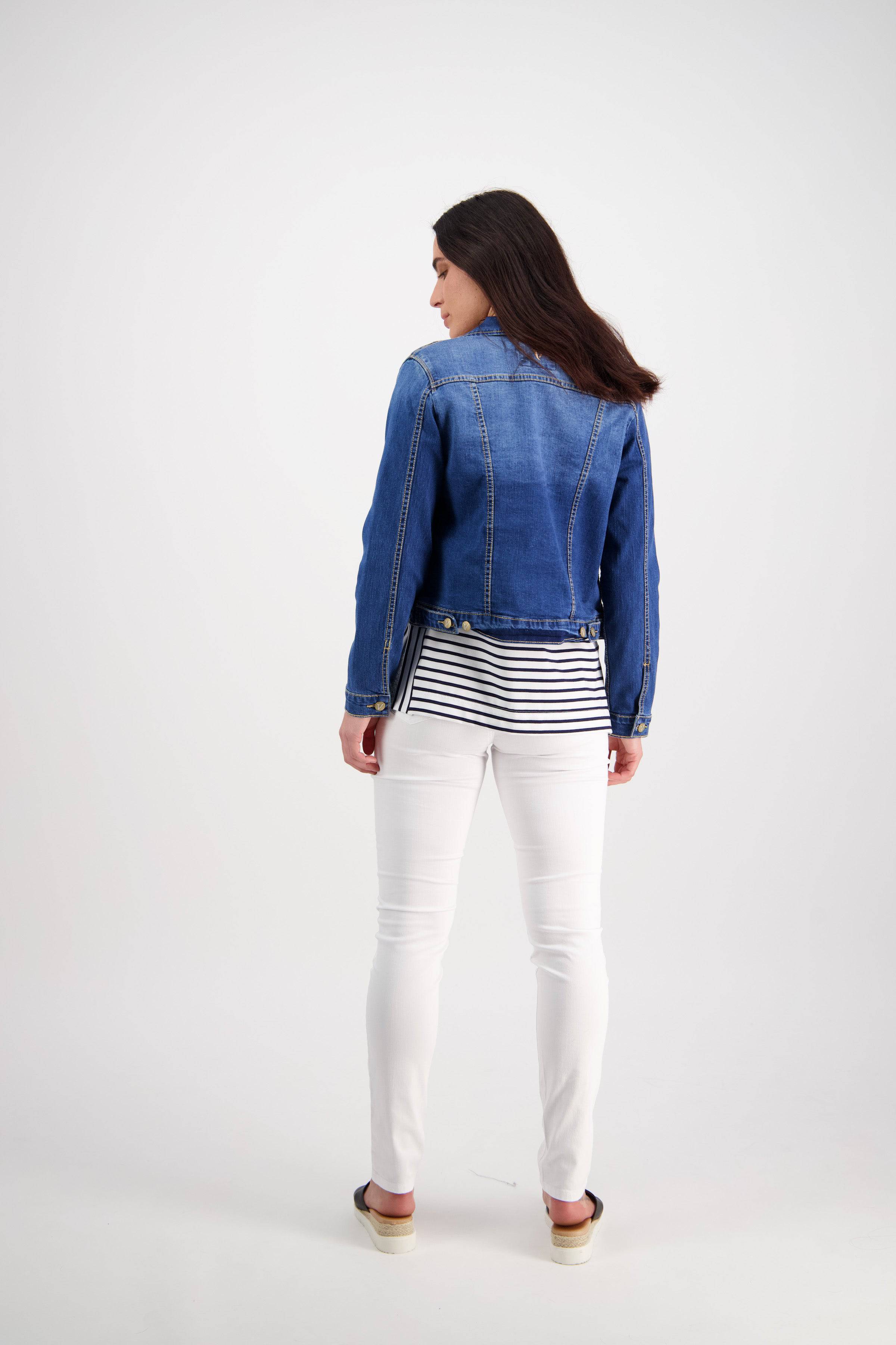 A woman faces away, wearing the Vassalli Ratio Jacket in blue wash denim with a black and white striped top, white pants, and sandals against a plain white background.