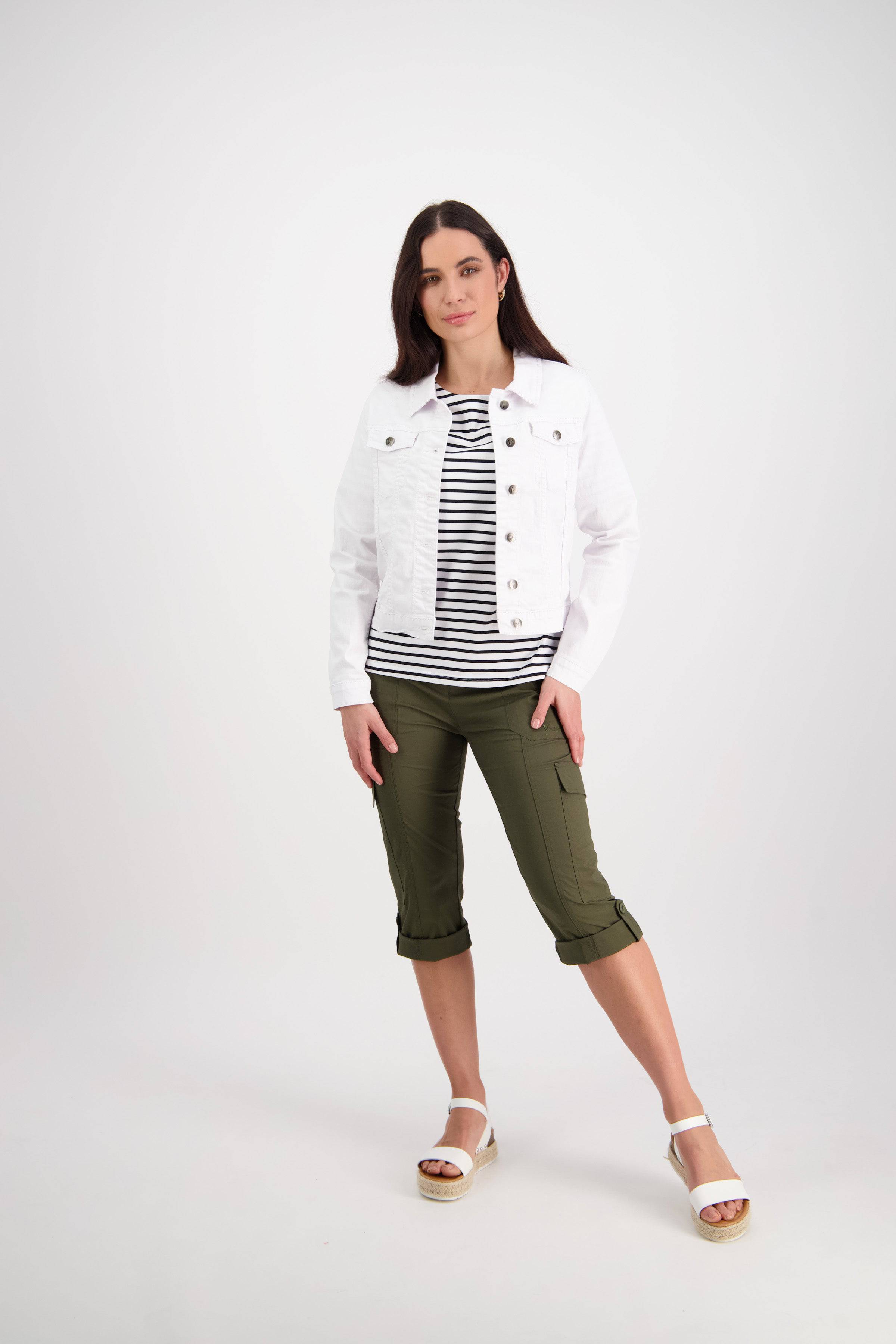 A woman with long dark hair stands confidently against a plain background, wearing the Vassalli Ratio Jacket in white denim over a black-and-white striped top, olive green capri pants, and white wedge sandals.