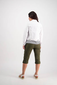 A woman with long dark hair stands facing away against a white background, wearing the Vassalli Ratio Jacket in white denim along with olive green capri pants and white sandals.