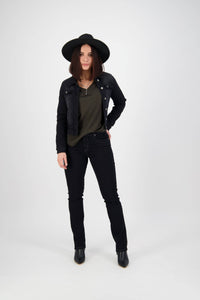 A woman stands against a plain white background, wearing the Vassalli Rider Knit Jacket in black, paired with black jeans and black ankle boots. She has one hand in her pocket and a relaxed expression.