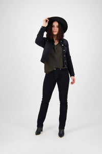 A woman stands against a plain white background, wearing the Vassalli Rider Knit Jacket in black, a wide-brim hat, dark green top, black jeans, and black ankle boots, holding her hat with one hand.