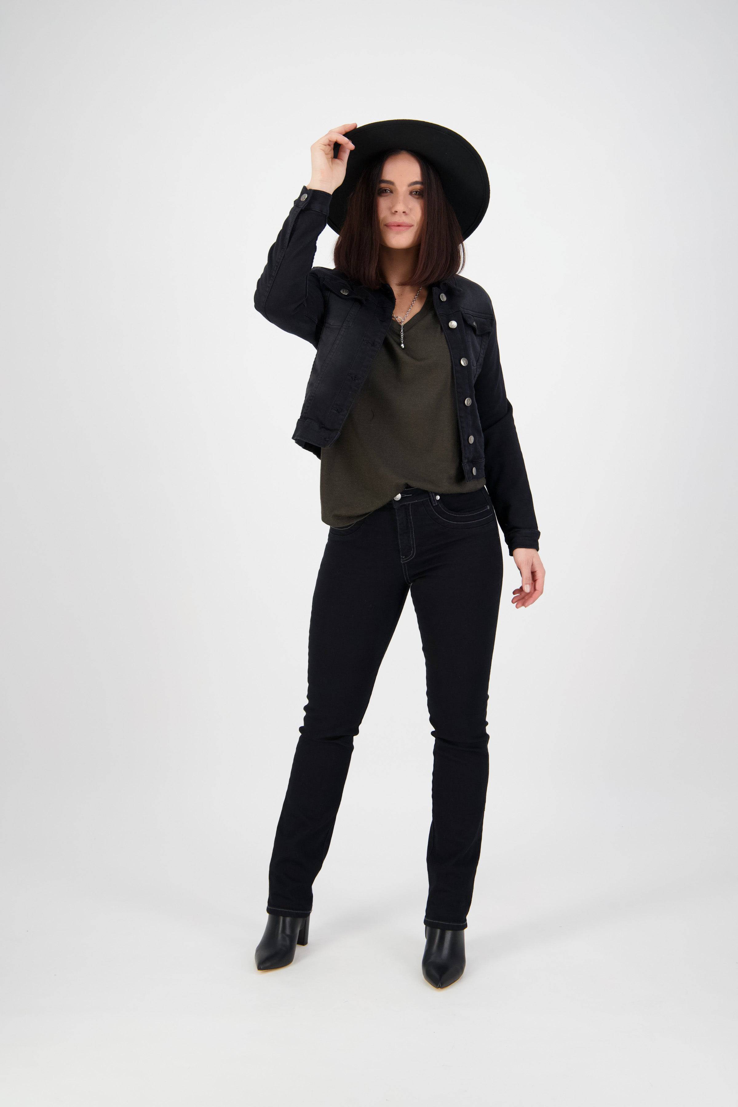 A woman stands against a plain white background, wearing the Vassalli Rider Knit Jacket in black, a wide-brim hat, dark green top, black jeans, and black ankle boots, holding her hat with one hand.