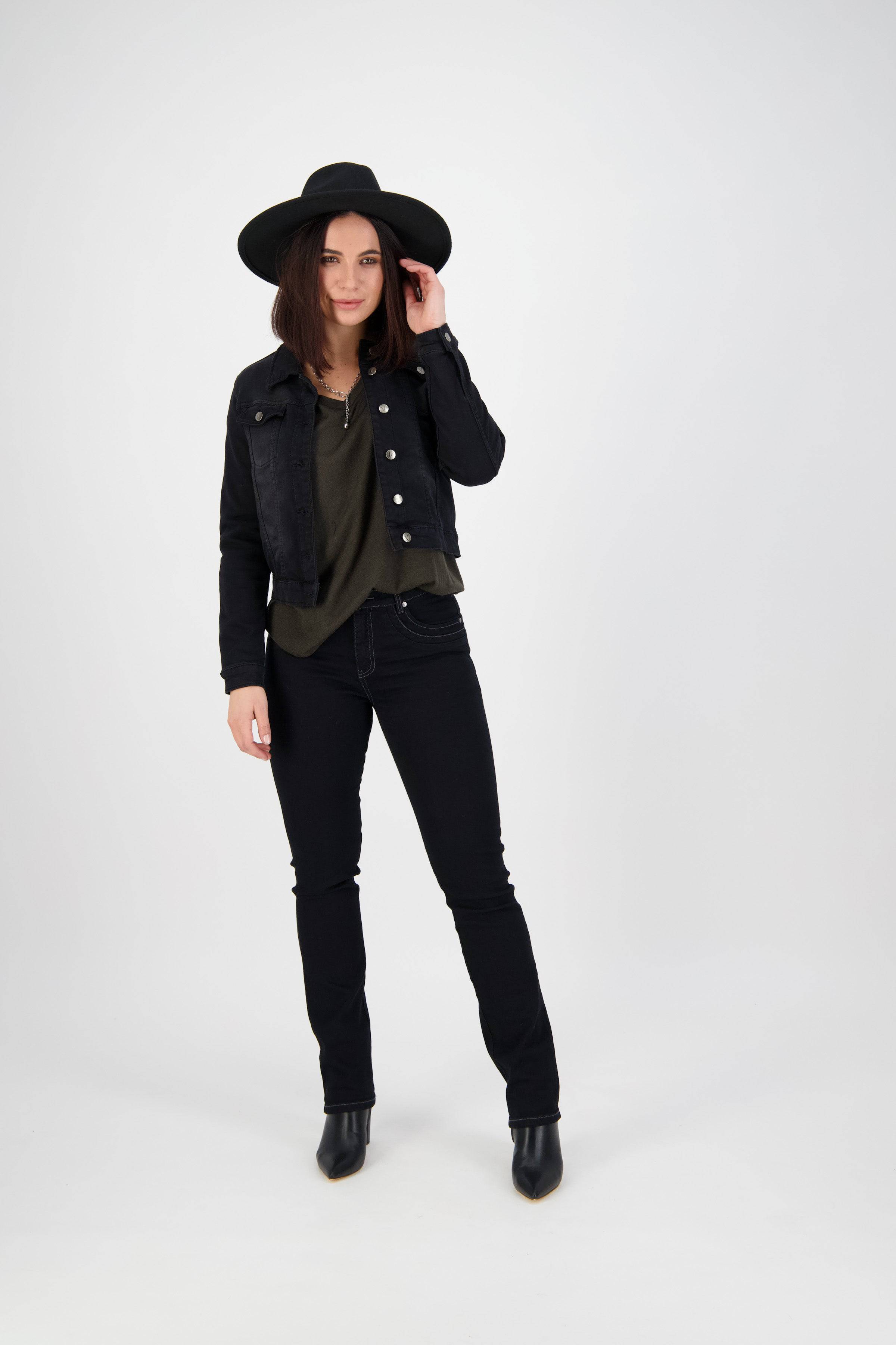 A woman stands against a white background, wearing the Vassalli Rider Knit Jacket in black, a black wide-brimmed hat, olive green top, black jeans, and black ankle boots. She touches her hat's brim and smiles slightly.