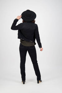A person stands facing away against a plain white background, wearing the Vassalli Rider Knit Jacket in black, dark jeans, heeled boots, and a black wide-brimmed hat, holding the brim with one hand.