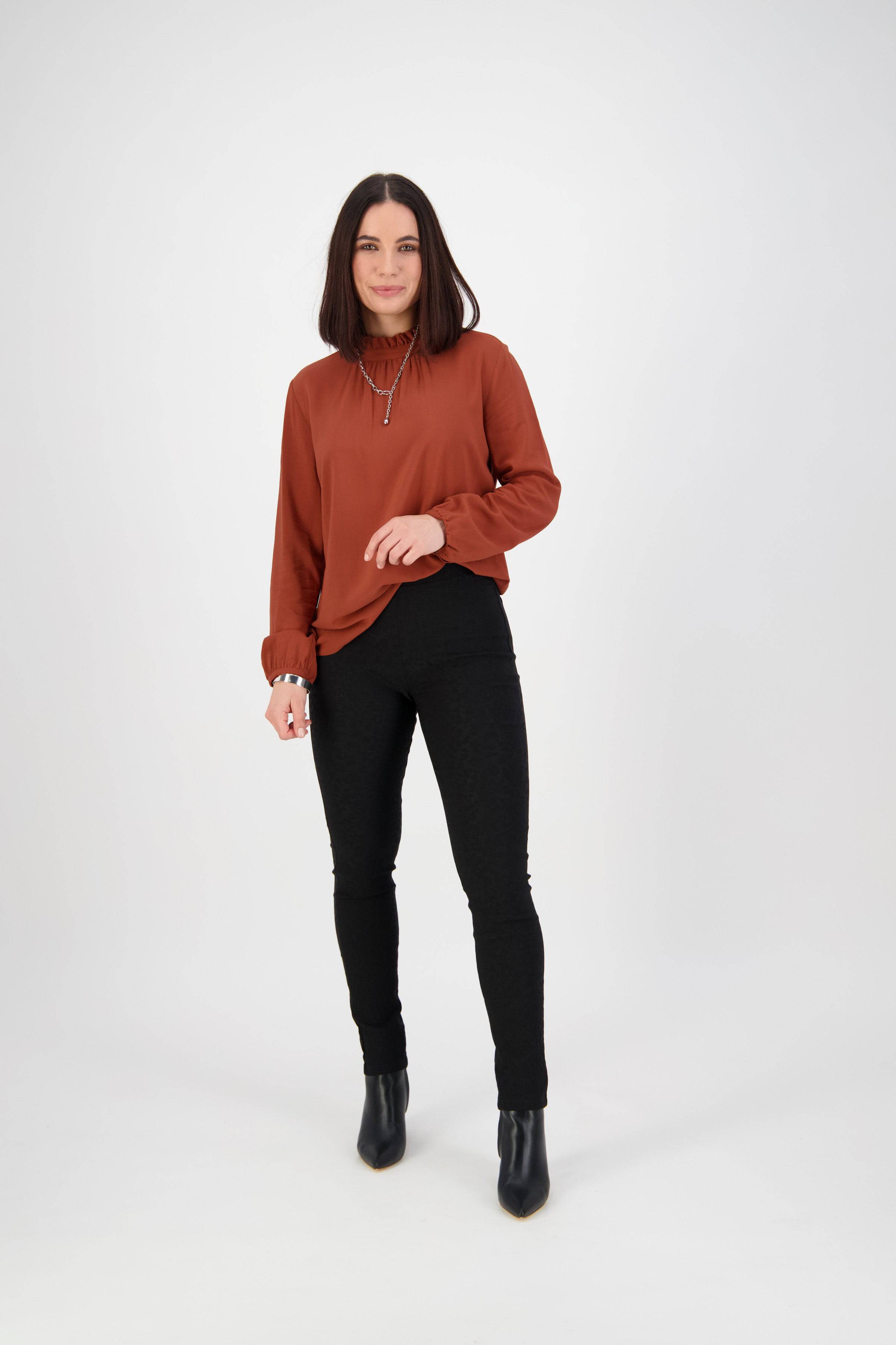 A woman with straight dark hair wearing a rust-colored long-sleeve top, Vassalli Origin Pull On - Black pants, and black ankle boots stands against a plain white background.