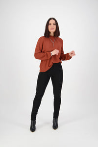 A woman with straight dark hair in a rust blouse, Vassalli Origin Pull On - Black pants, and ankle boots stands against a plain white background, smiling slightly and posing confidently.