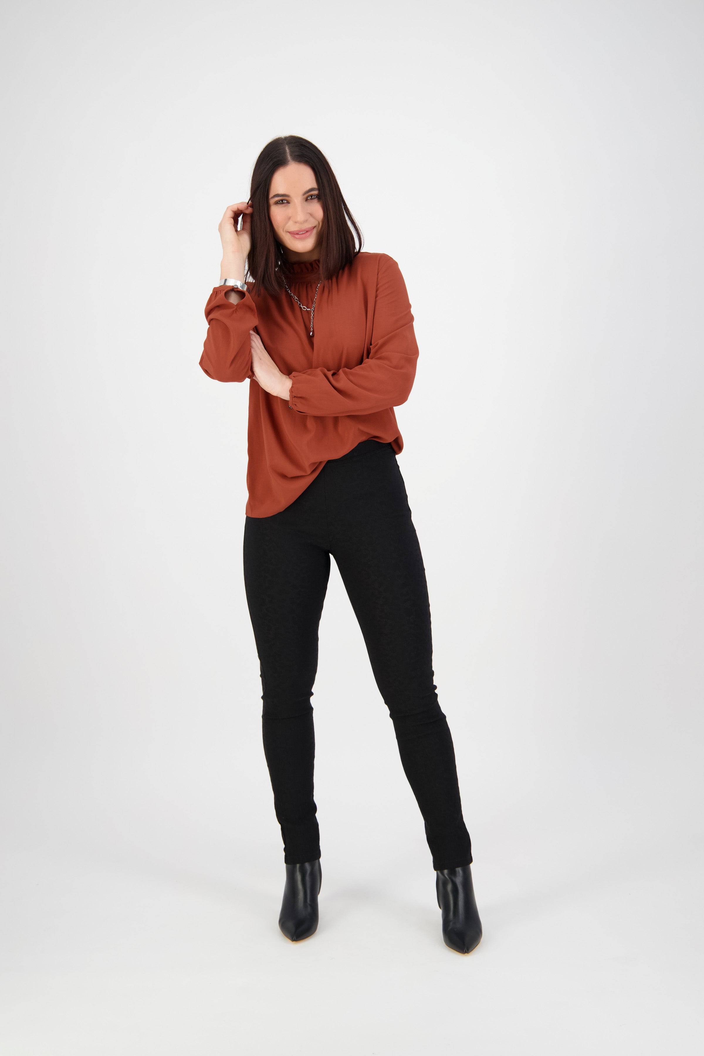 A woman with straight brown hair, wearing a rust blouse, Vassalli Origin Pull On - Black pants, and black boots stands against a plain white background, smiling with one hand touching her hair.