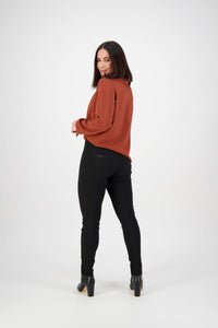 A woman with straight dark hair faces away, looking over her shoulder, wearing a rust blouse, Vassalli Origin Pull On - Black pants, and heeled boots against a plain white background.