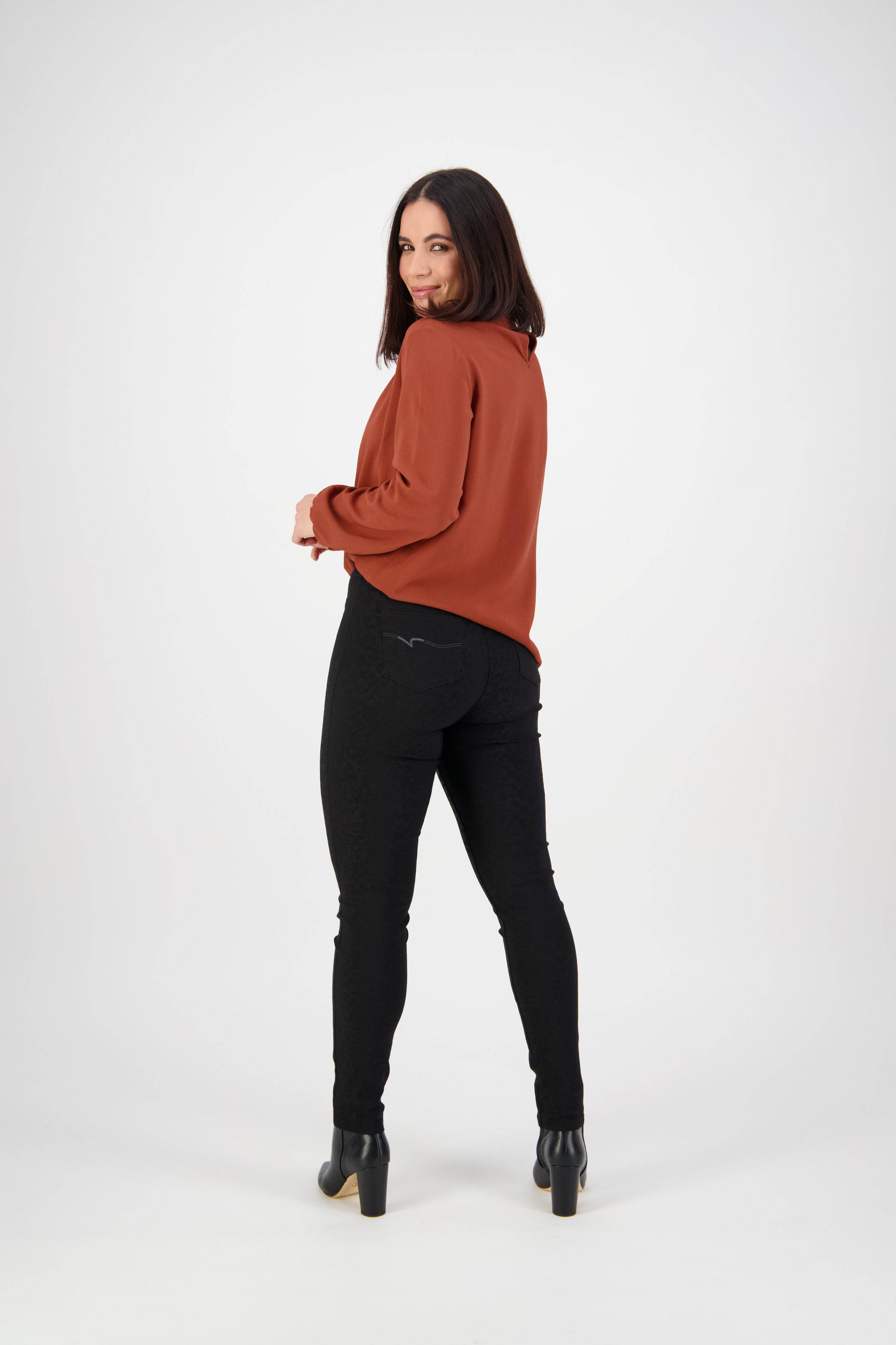 A woman with straight dark hair faces away, looking over her shoulder, wearing a rust blouse, Vassalli Origin Pull On - Black pants, and heeled boots against a plain white background.