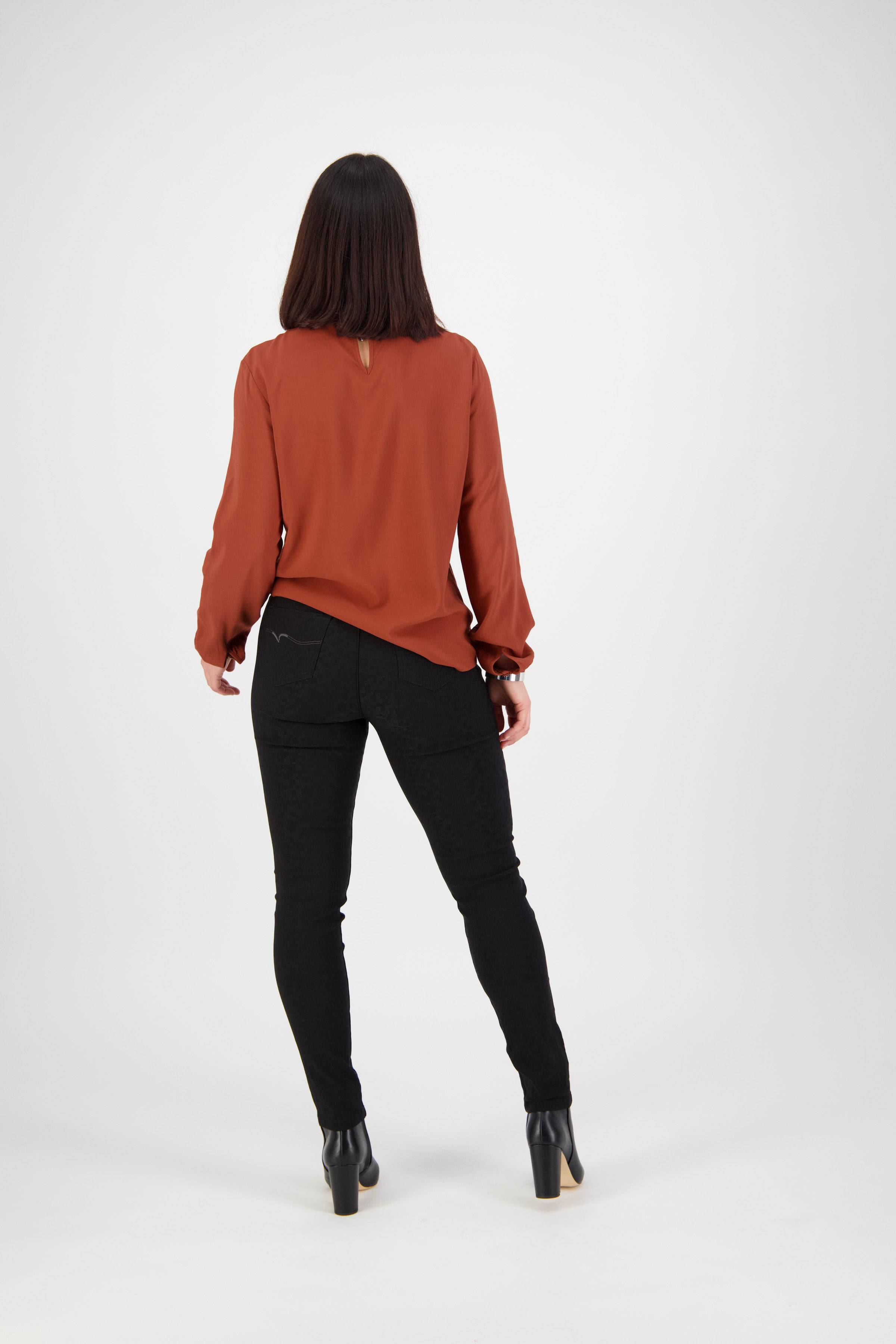 A woman with shoulder-length dark hair, facing away from the camera, wears a rust long-sleeve blouse and Vassalli Origin Pull On - Black pants with black heeled boots, standing against a plain white background.