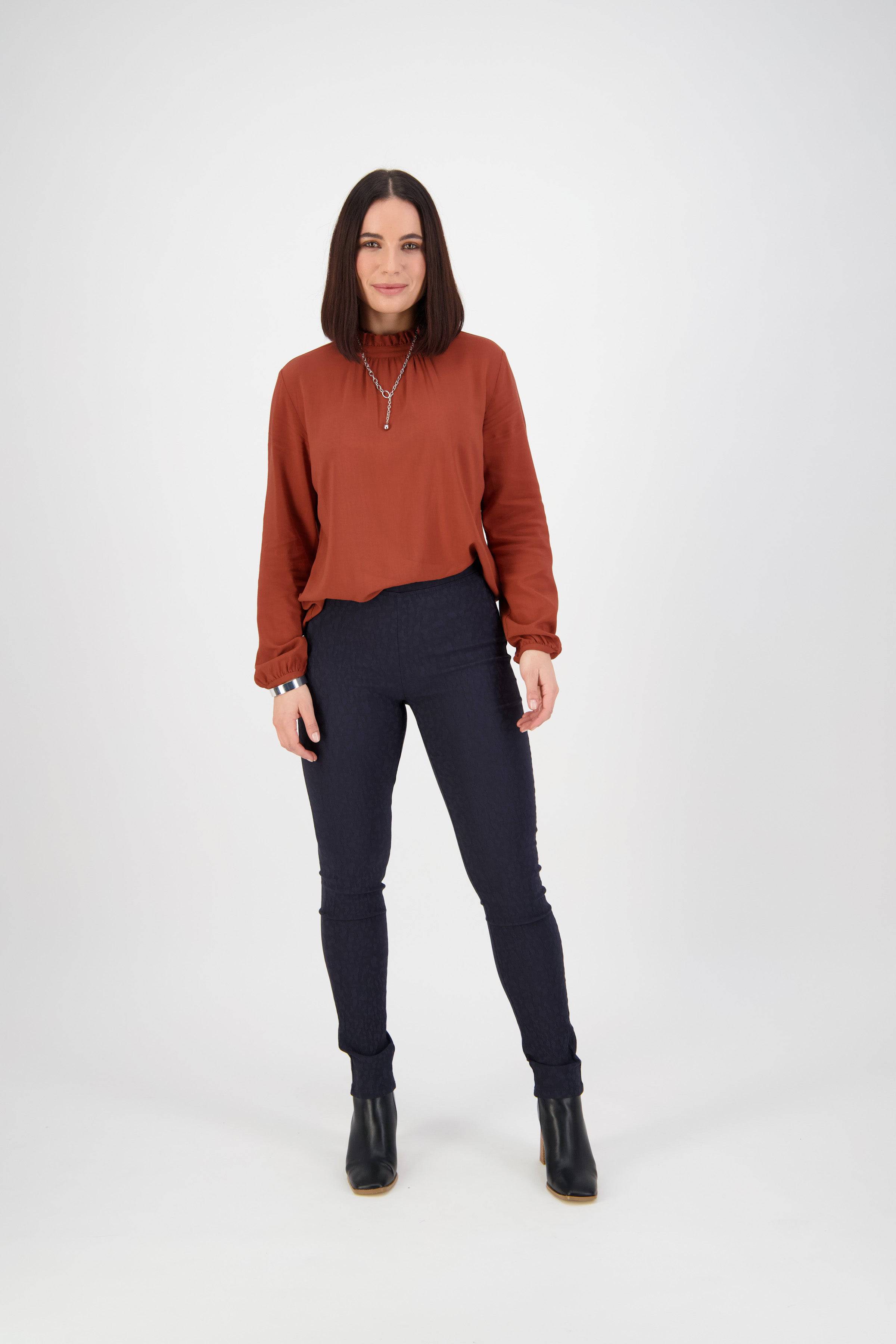 A woman with straight dark hair faces forward, wearing a rust-colored long-sleeve blouse, Vassalli Origin Pull On - Navy skinny leg stretch pants, and black ankle boots against a plain light background.