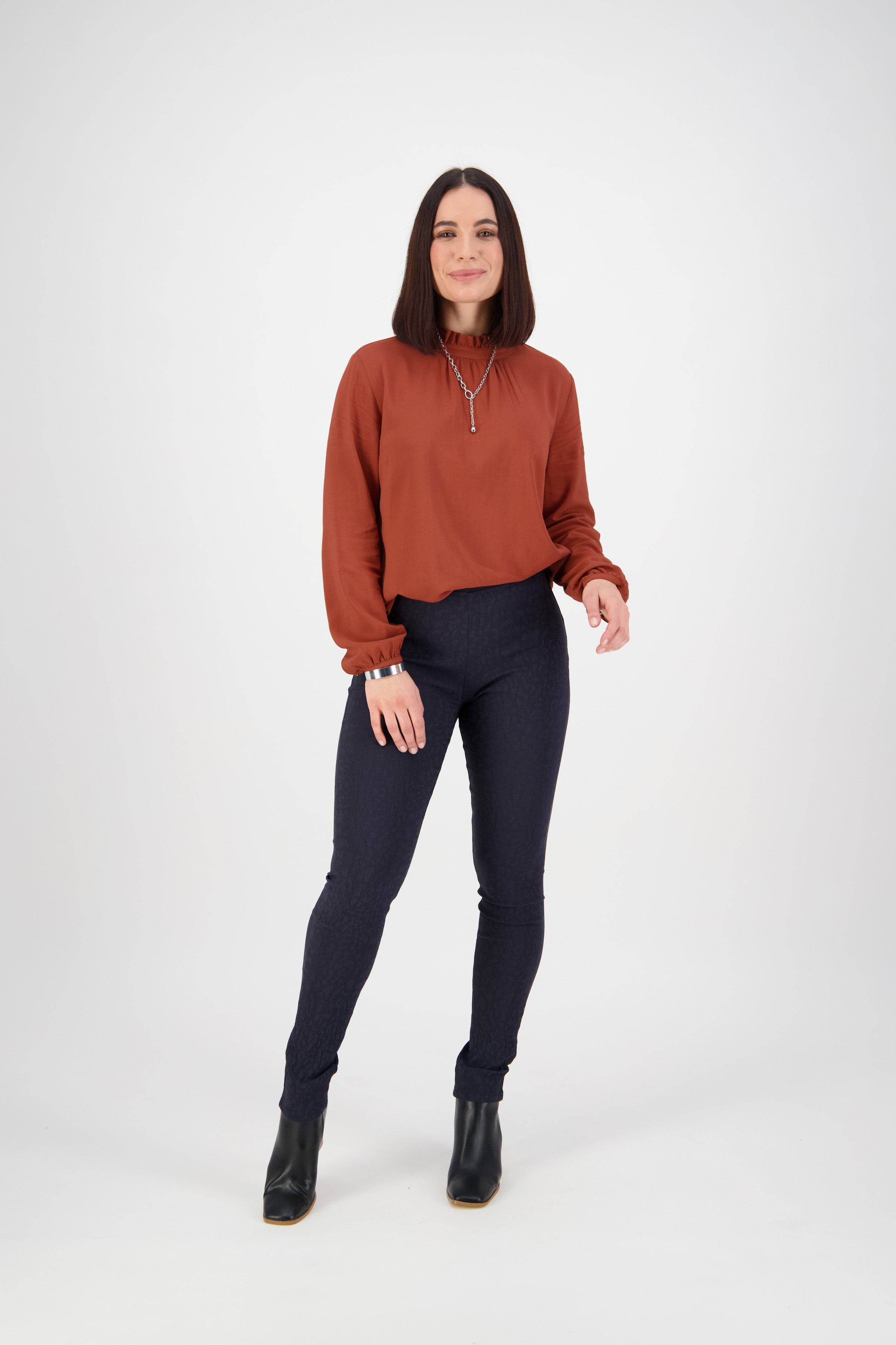 A woman with straight, shoulder-length dark hair stands against a white background wearing a rust-colored long-sleeve blouse, Vassalli Origin Pull On - Navy skinny pants, black ankle boots, and is smiling slightly.