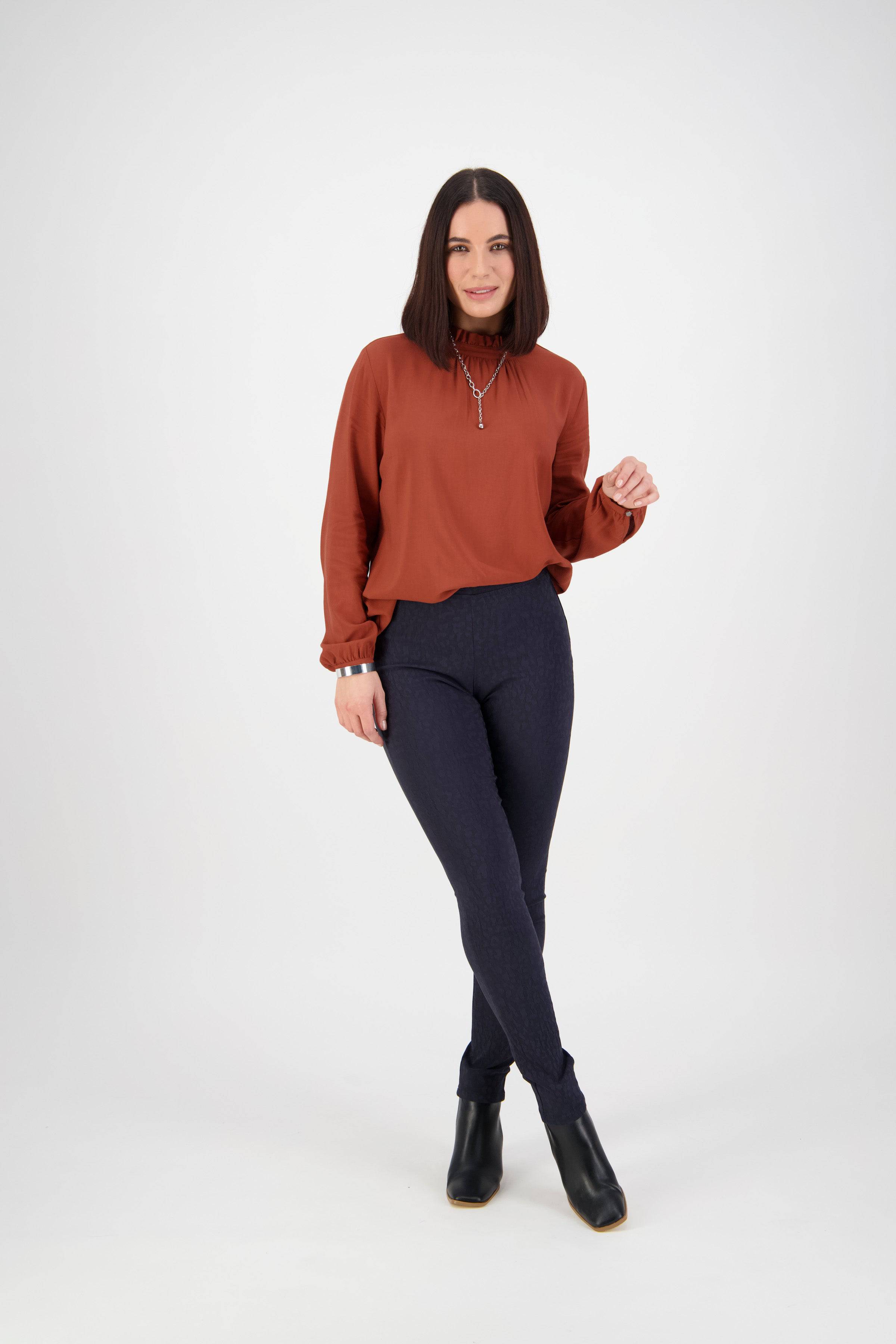 A woman with straight dark hair stands against a white background, wearing the Vassalli Origin Pull On - Navy pants and a rust long-sleeve blouse with black ankle boots. She smiles slightly, holding one hand near her face.