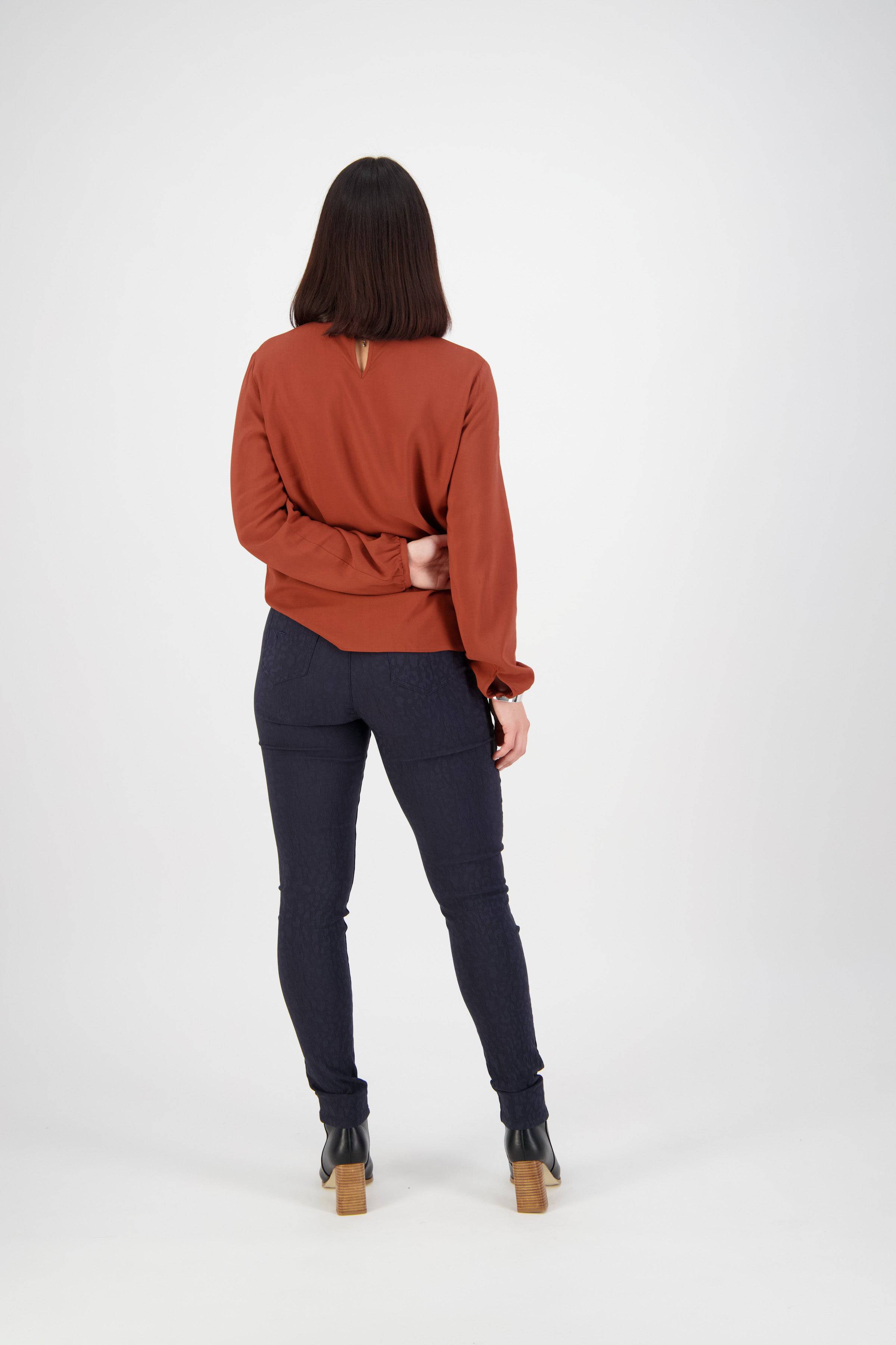 A person with straight, shoulder-length brown hair stands with their back to the camera, wearing a rust sweater, Vassalli Origin Pull On - Navy pants, and black heeled boots against a plain white background.