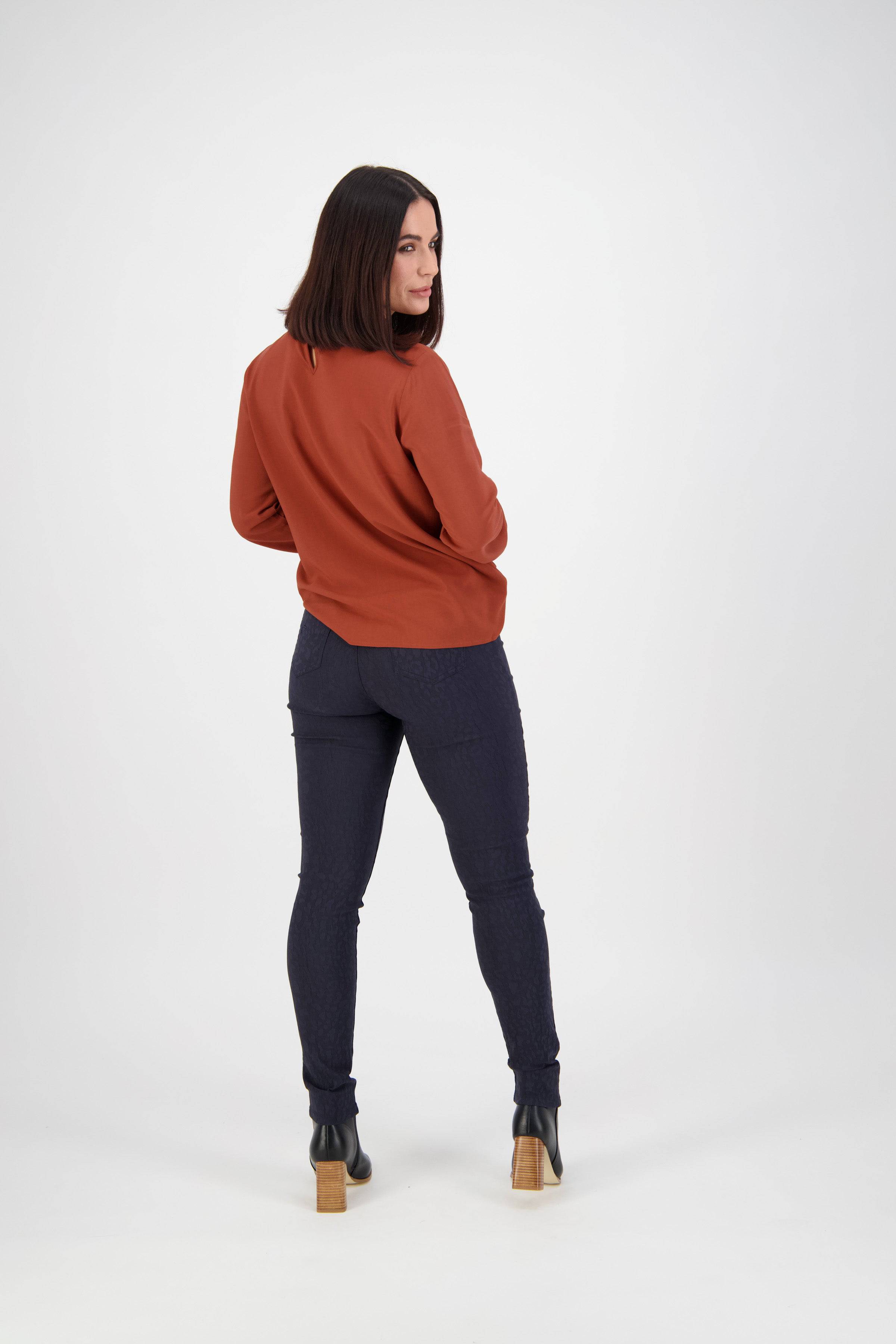 A person with shoulder-length dark hair wears a rust long-sleeve top and Vassalli Origin Pull On - Navy skinny jeans, paired with black heeled ankle boots, standing against a plain white background and looking over their shoulder.