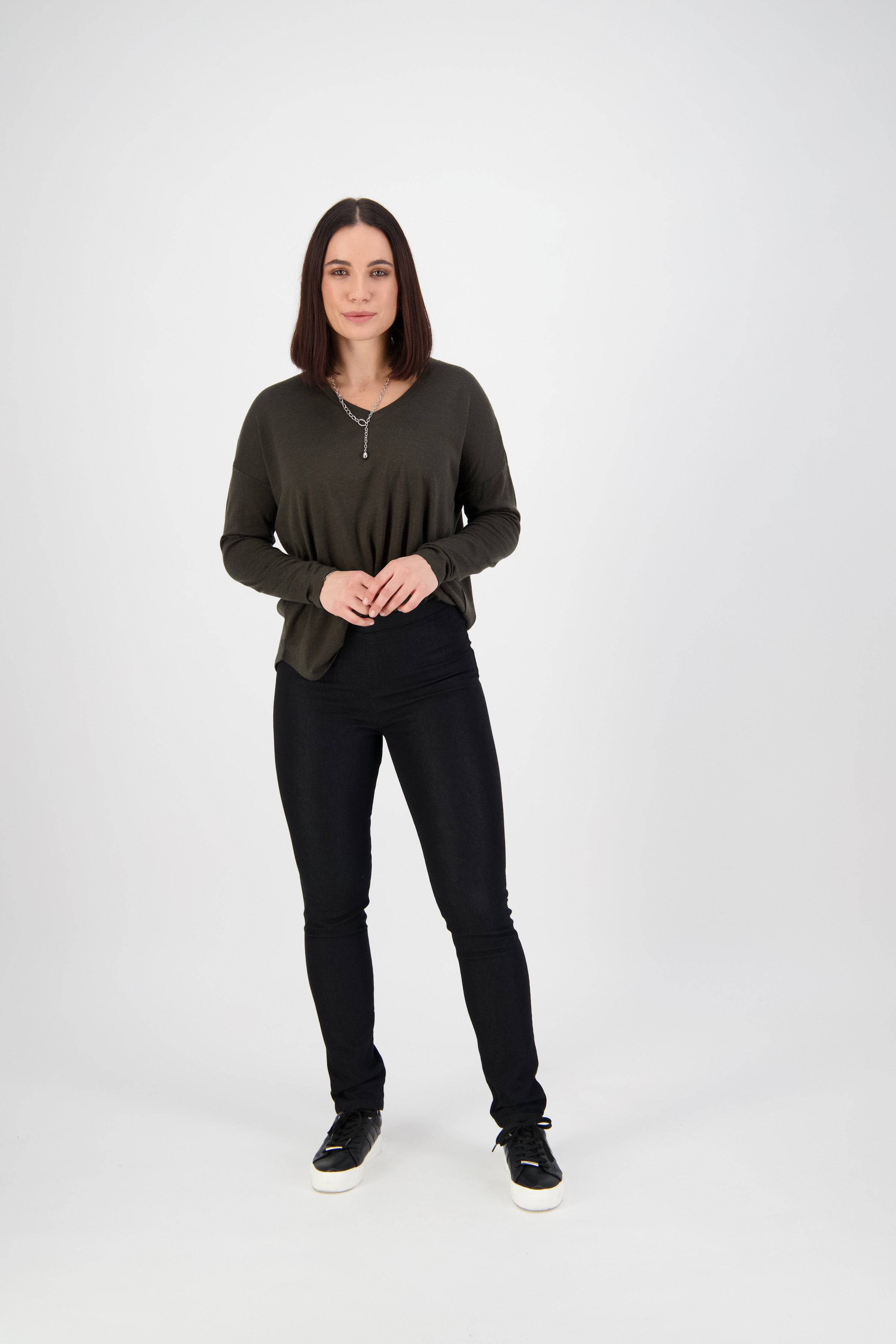 A woman with straight brown hair stands against a white background, wearing a dark green long-sleeve top, Vassalli Origin Pull On - Black Denim pants, and black sneakers, facing forward with her hands clasped.