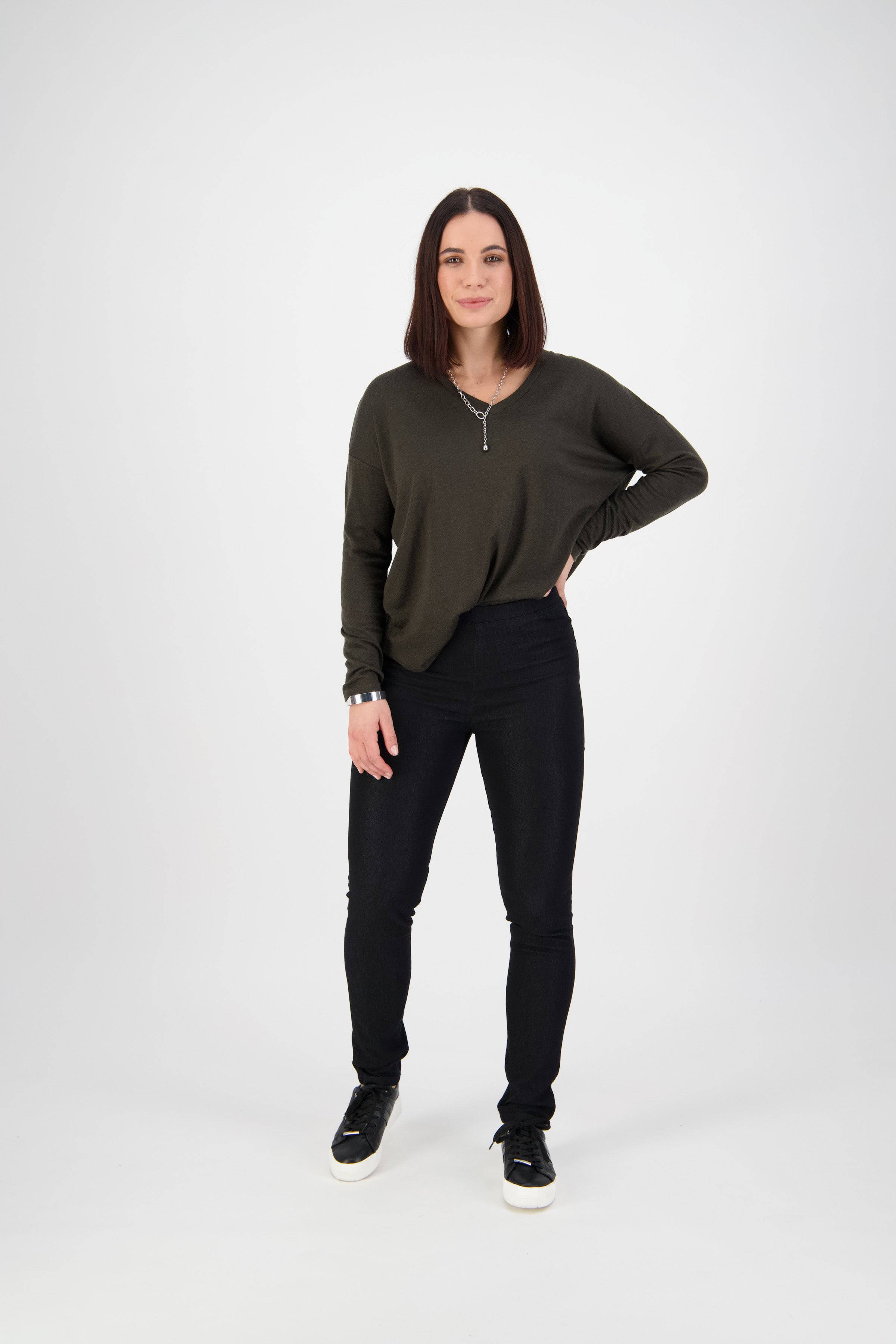 A woman with straight, shoulder-length dark hair stands against a white background, wearing a dark green long-sleeve top, Vassalli Origin Pull On - Black Denim pants, and black sneakers. She rests one hand on her hip and smiles slightly.