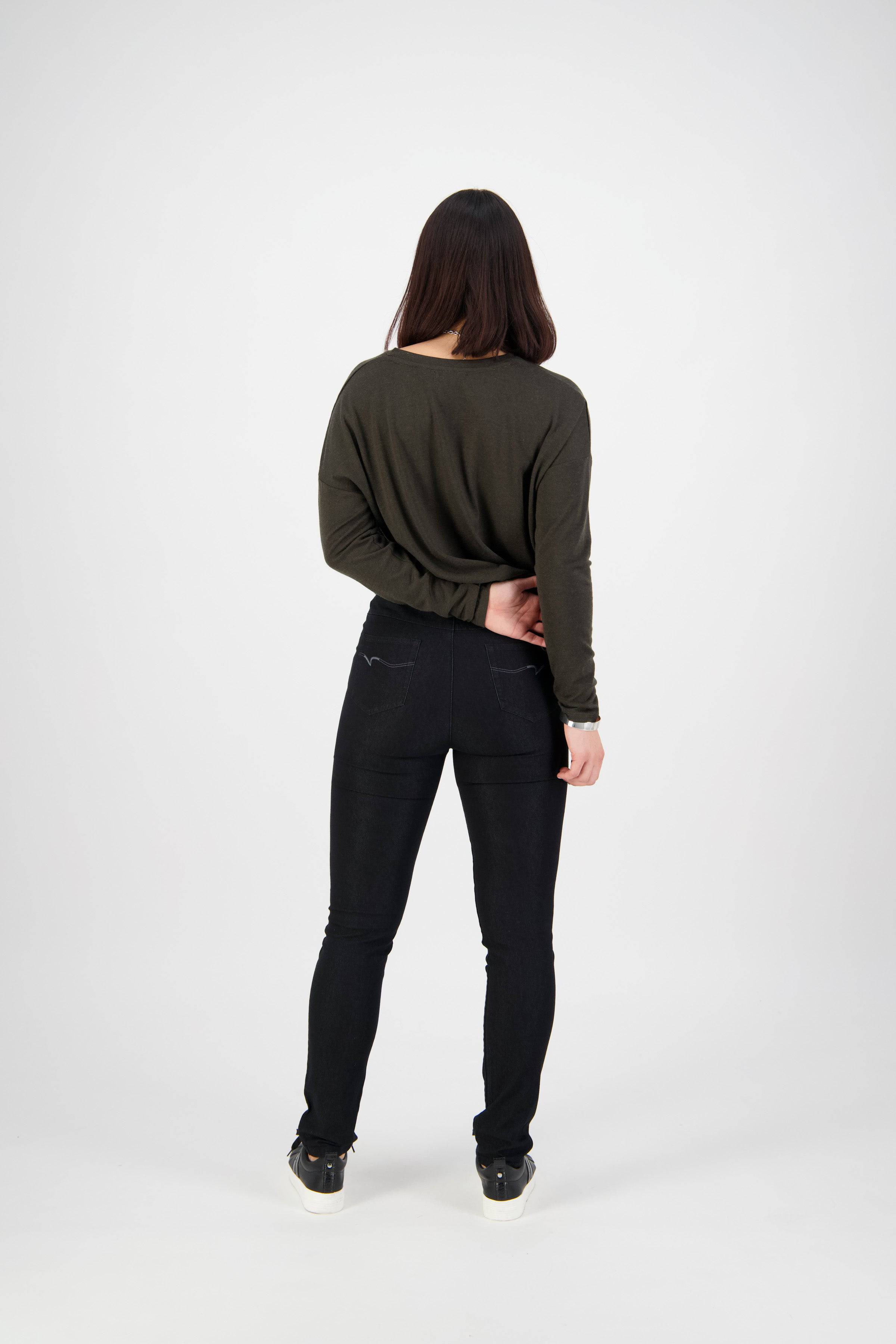 A woman with straight dark hair, facing away on a plain white background, wears a dark green long-sleeve shirt, sneakers, and Vassalli Origin Pull On - Black Denim skinny leg pants. One arm is behind her lower back.