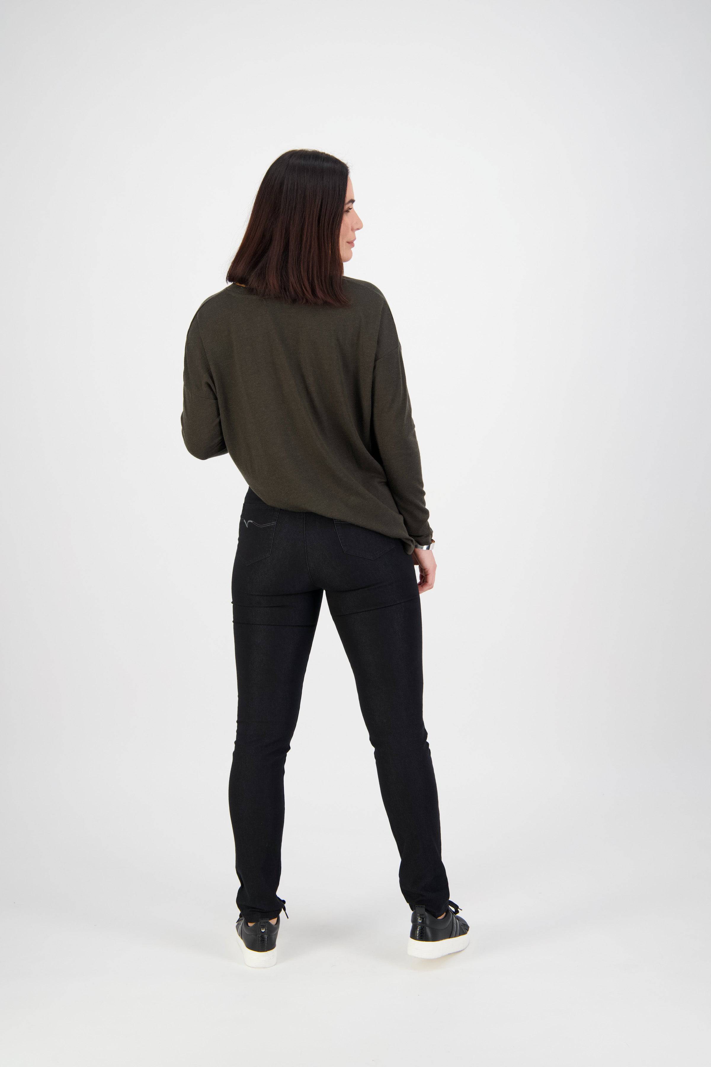 A woman with straight dark hair, wearing a long-sleeve olive green top, Vassalli Origin Pull On - Black Denim, and black sneakers, stands facing away from the camera against a plain white background.