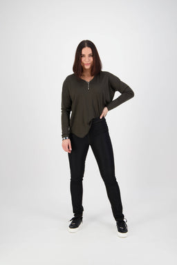 A woman with straight, shoulder-length brown hair stands confidently against a white background, wearing Vassalli's Origin Pull On - Black Denim (Contrast Stitch) pants, a dark green top, and black sneakers, smiling with one hand on her hip.
