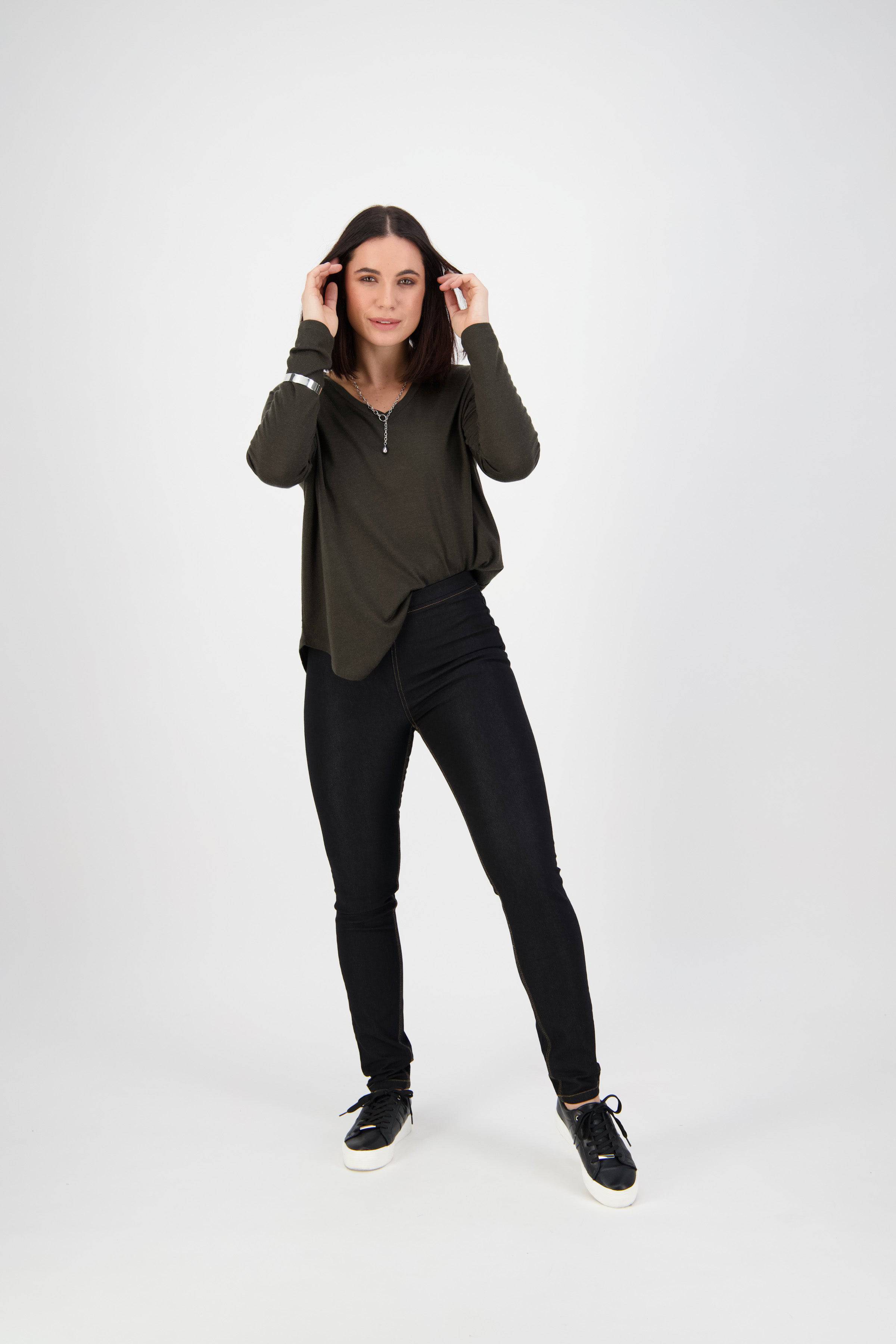 A woman with straight dark hair poses against a white background, wearing Vassalli's Origin Pull On - Black Denim (Contrast Stitch) pants, a dark green long-sleeve top, and black sneakers, hands near her head and looking at the camera.