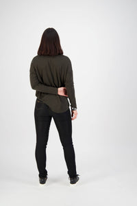 A person with shoulder-length dark hair wears a dark long-sleeve top and Vassalli Origin Pull On - Black Denim (Contrast Stitch) pants, standing facing away with one arm bent behind their back against a plain white background.