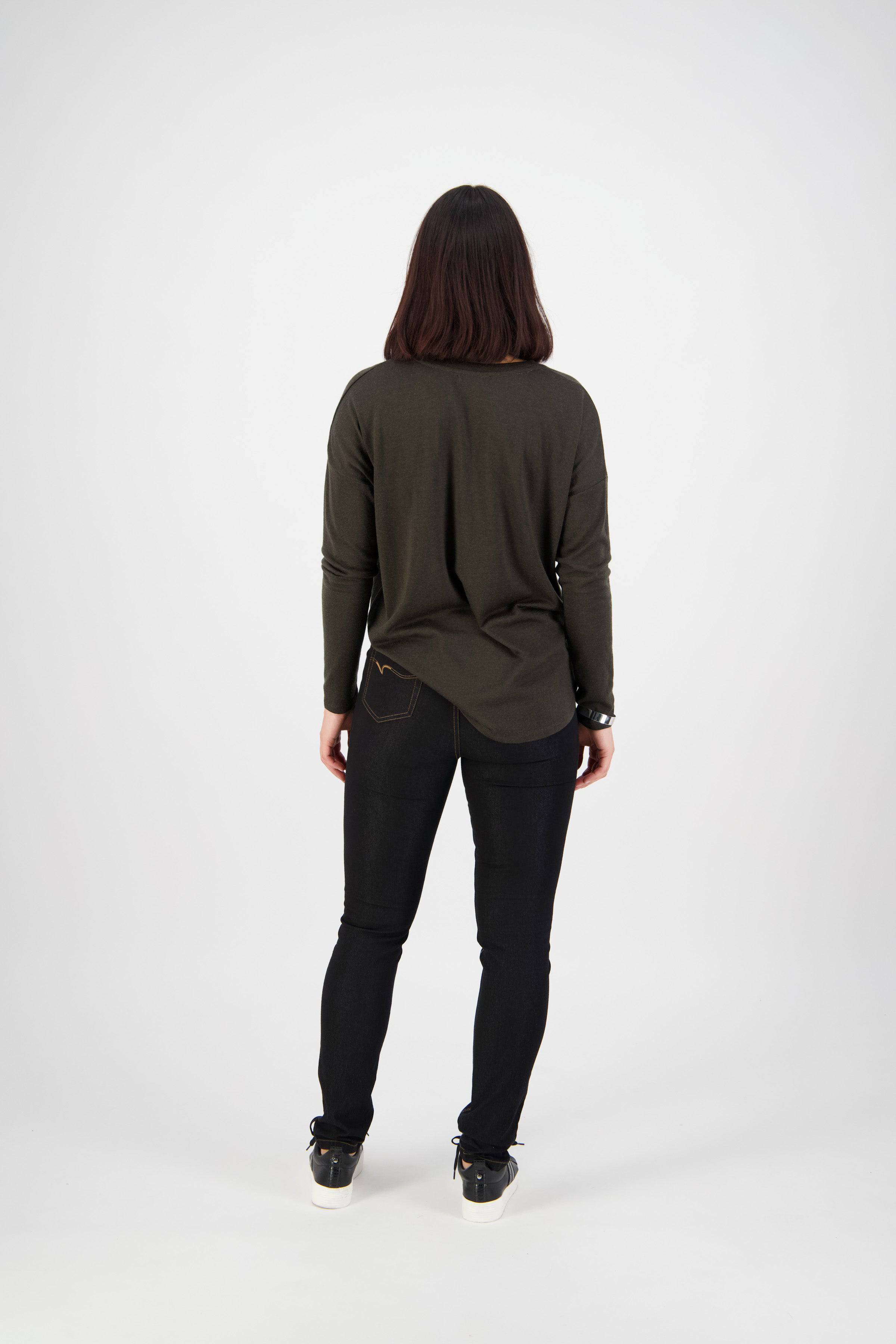 A person with shoulder-length dark hair, facing away, wears a long-sleeved dark green top and Vassalli's Origin Pull On - Black Denim (Contrast Stitch) pants with black sneakers, set against a plain white background.