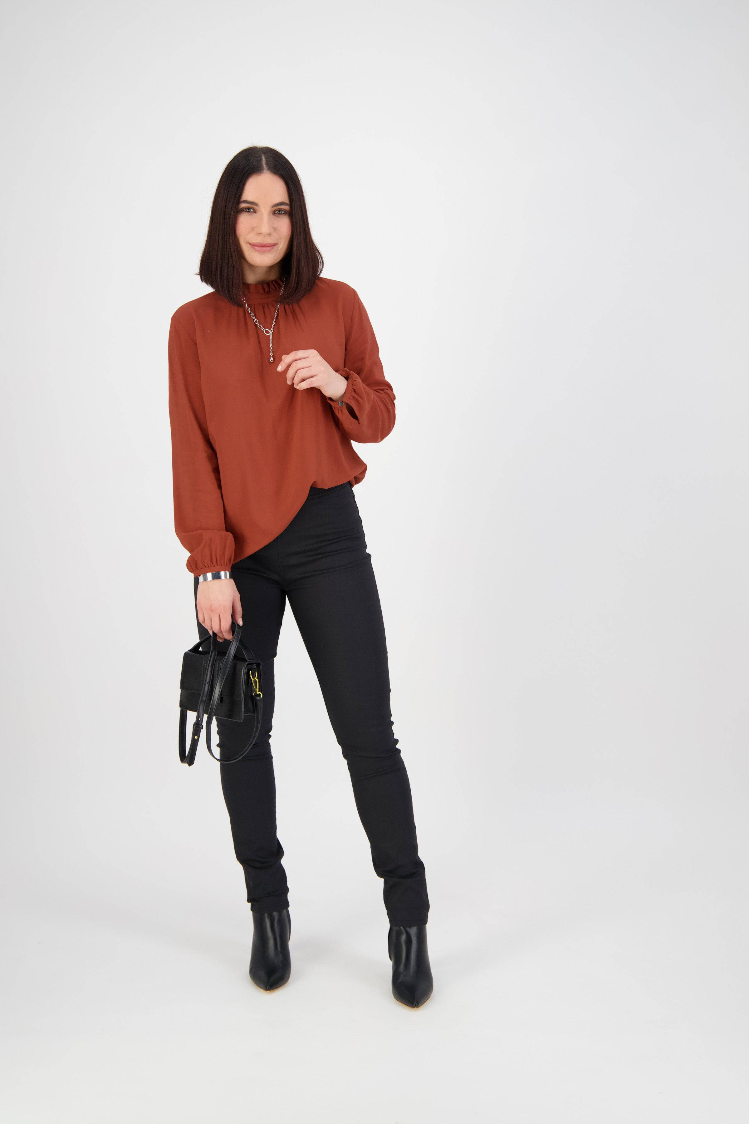 A woman with shoulder-length dark hair wears a rust-colored blouse and Vassalli's Origin Pull On - Black pants, paired with black boots and a black handbag. She stands against a plain white background, smiling slightly.