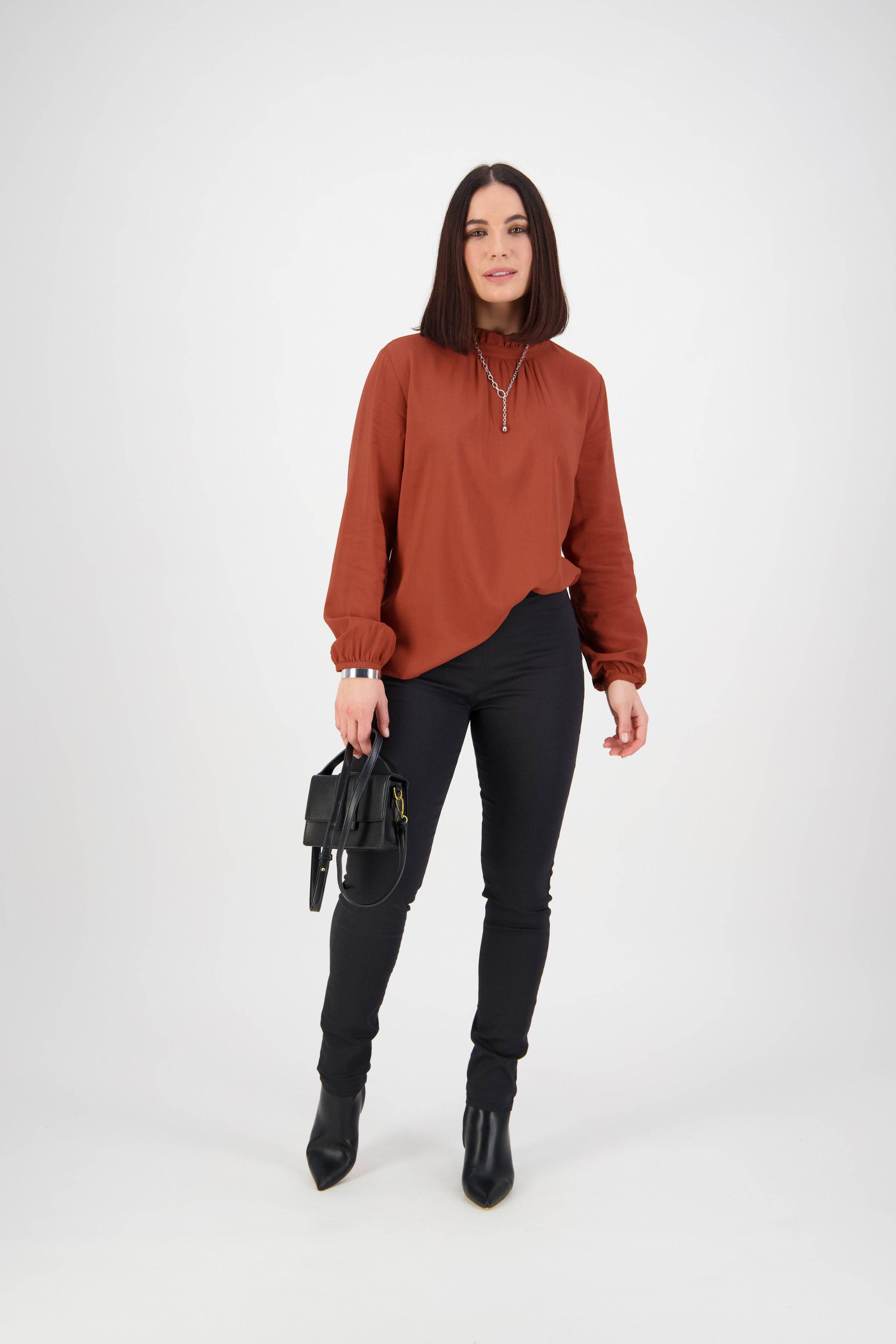 A woman with straight dark hair wears a rust blouse, Vassalli Origin Pull On - Black pants, and black ankle boots. She stands against a plain white background, holding a small black handbag in her left hand.
