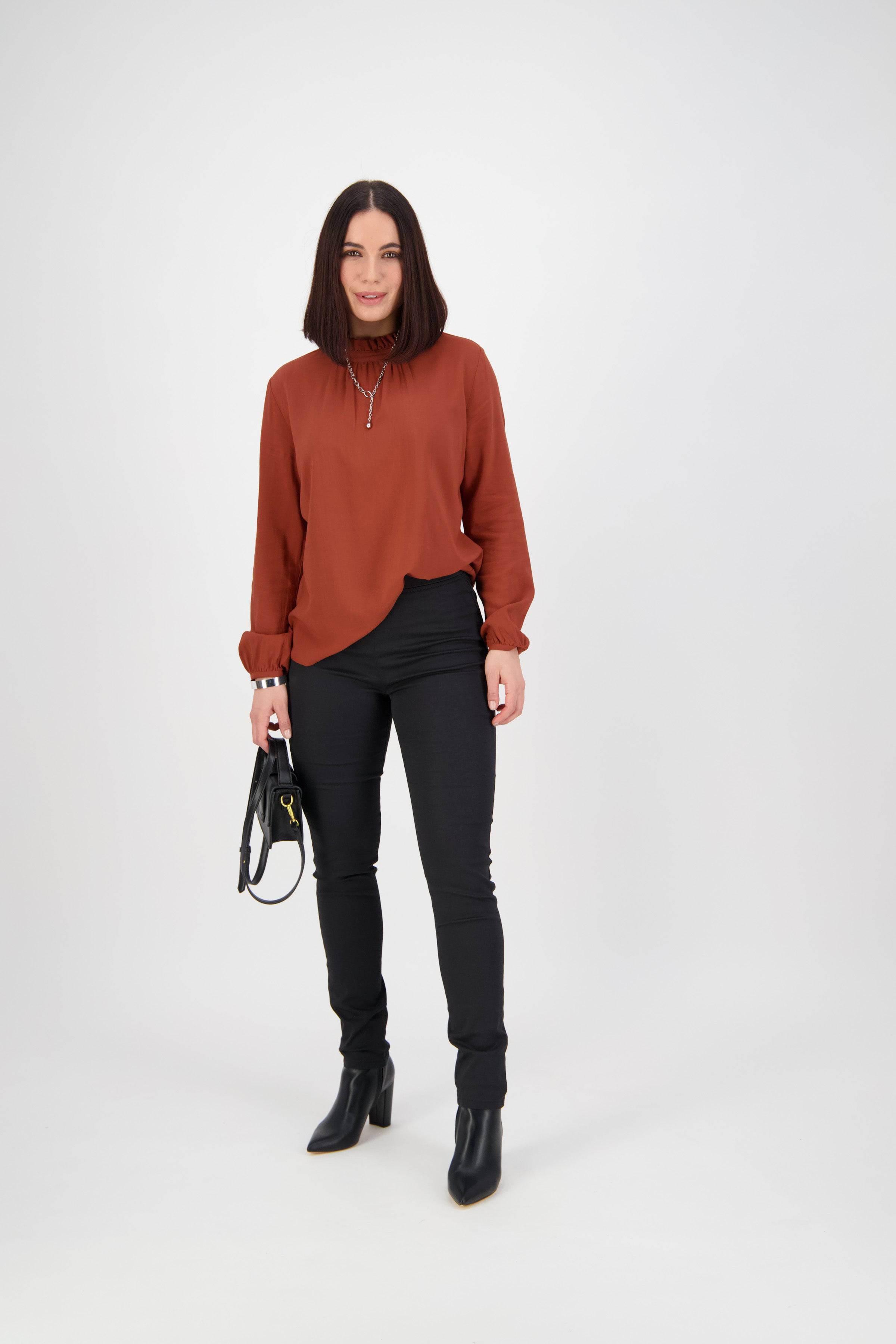 A woman with straight dark hair wears a rust blouse, Vassalli Origin Pull On - Black pants, and black ankle boots, holding a black handbag in a plain white studio.
