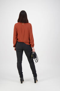 A woman with straight dark hair, facing away, wears a rust long-sleeve blouse and Vassalli Origin Pull On - Black skinny leg pants, holding a black handbag in her left hand against a plain white background.