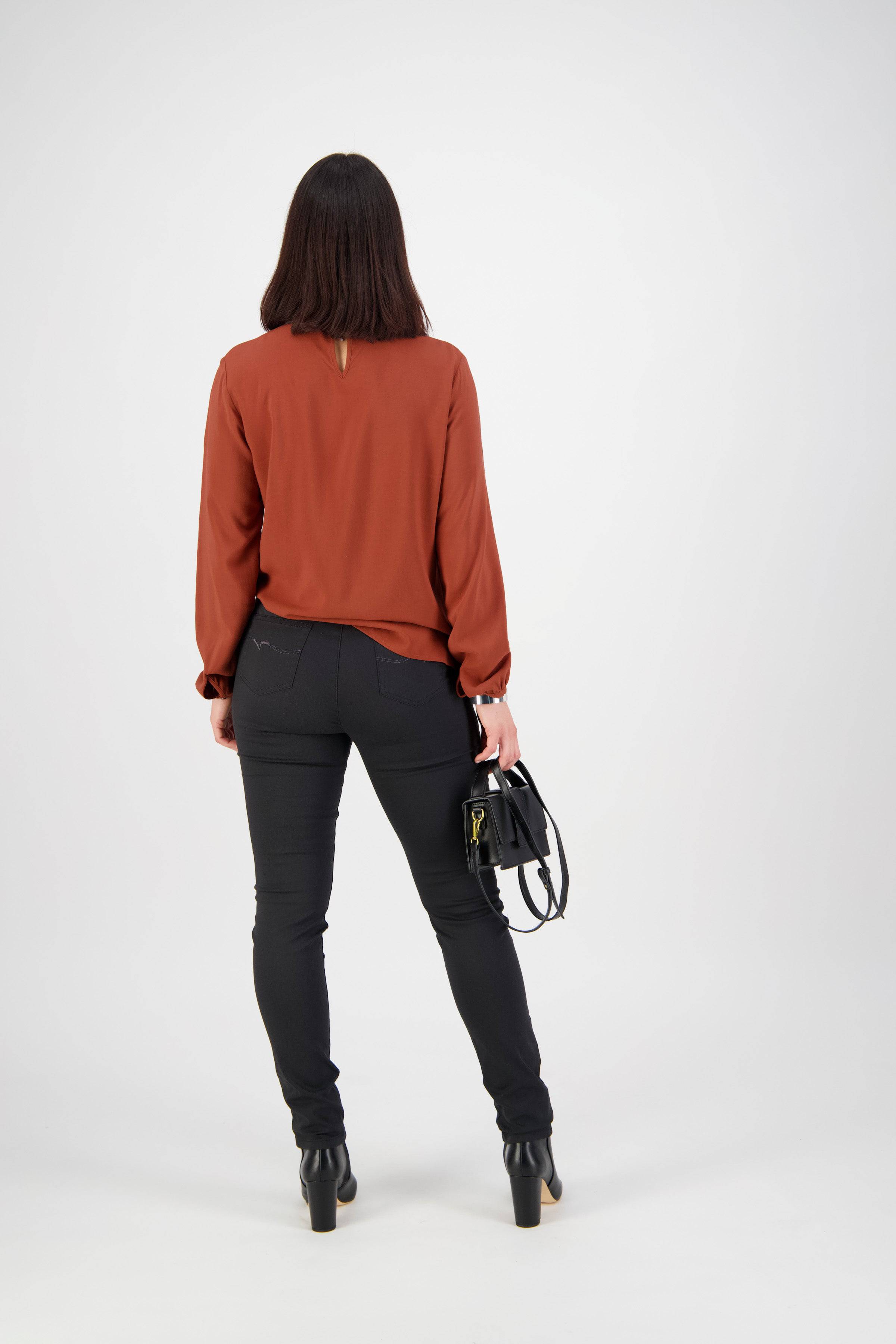 A woman with straight dark hair, facing away, wears a rust long-sleeve blouse and Vassalli Origin Pull On - Black skinny leg pants, holding a black handbag in her left hand against a plain white background.