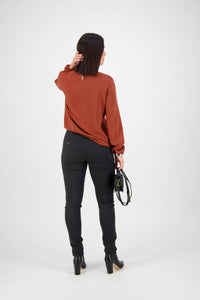 A woman with straight dark hair faces away, wearing a rust long-sleeve blouse and Vassalli Origin Pull On - Black pants, paired with black heeled boots and a black handbag, set against a plain white background.