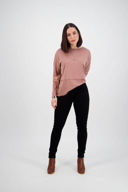 A woman with straight brown hair stands against a white background, wearing a pink long-sleeve top, Vassalli Origin Cord Pull On pants in black, brown boots, and a necklace, one hand behind her back and a slight smile.