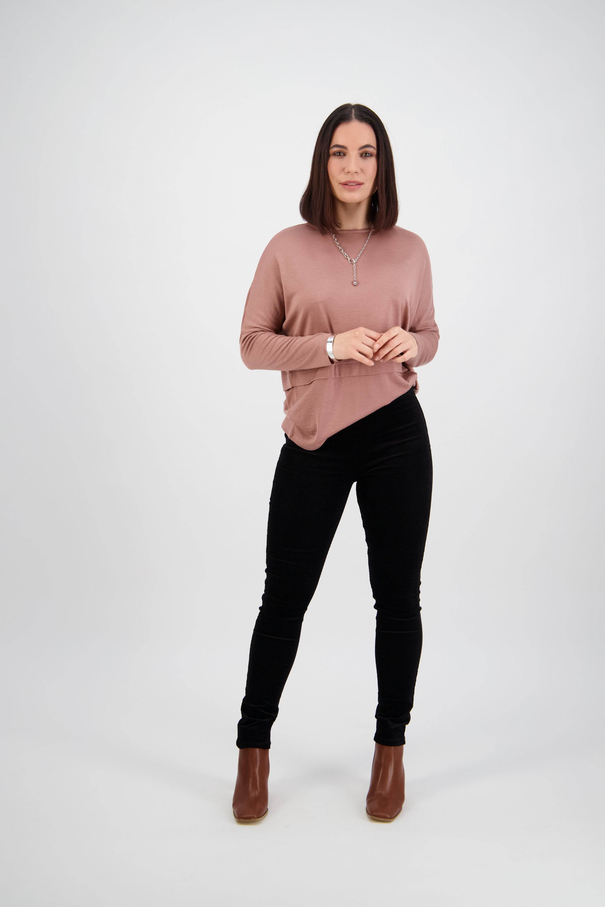 A woman with straight dark hair wears a mauve sweater, Vassalli Origin Cord Pull On - Black pants, and brown ankle boots, standing against a plain white background. She faces the camera neutrally, hands loosely clasped.