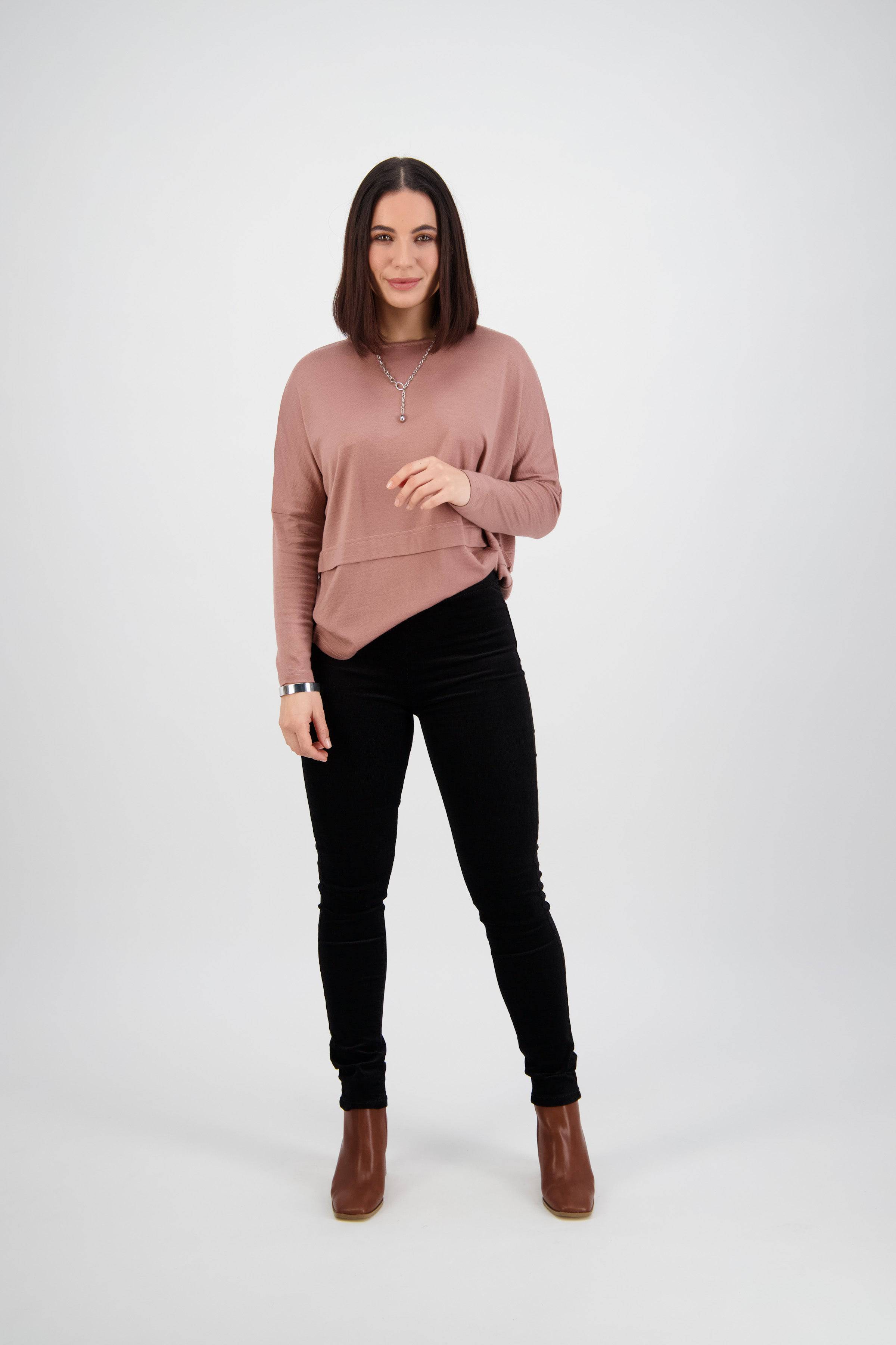 A woman with straight, shoulder-length brown hair stands against a white background, wearing a pink long-sleeve top, Vassalli Origin Cord Pull On pants in black, and brown ankle boots. She is smiling slightly and holding a necklace.
